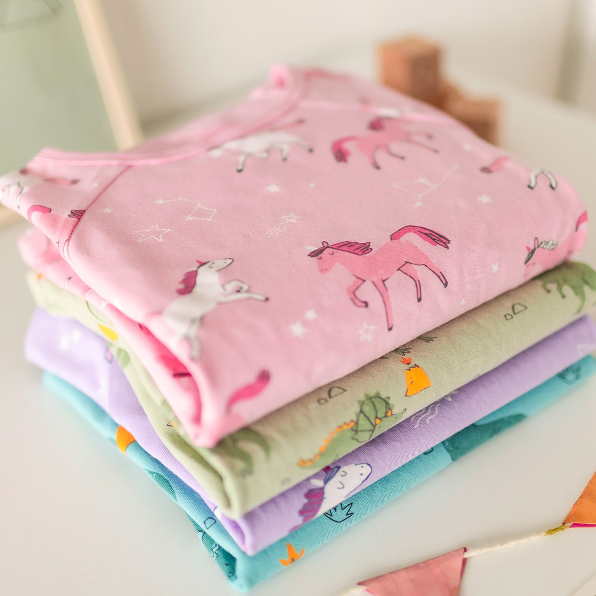 Pile de pyjamas Souris Mini pour enfants, avec un modèle rose à licornes au-dessus et d’autres aux motifs dinosaures en dessous.