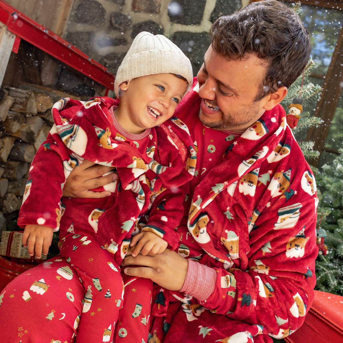 Papa et enfant en pyjamas et peignoirs rouges à motifs de Noël partageant un moment complice et chaleureux près du sapin.