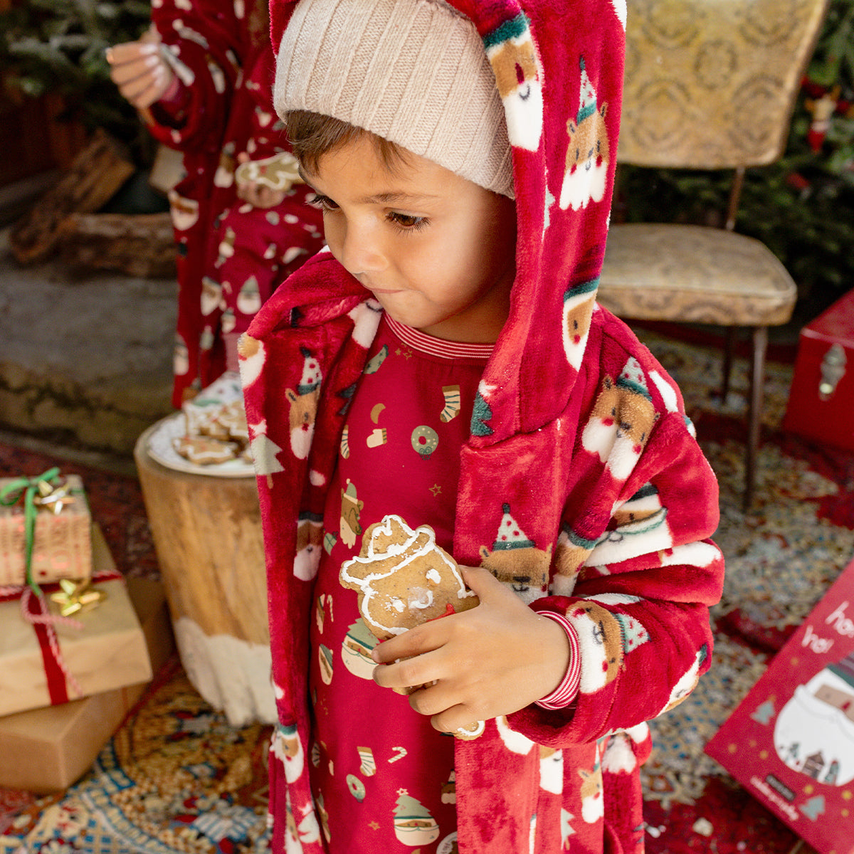Enfant en robe de chambre rouge à motifs de Noël tenant un biscuit pain d’épice, vêtu d’un pyjama assorti pour un moment festif et douillet.