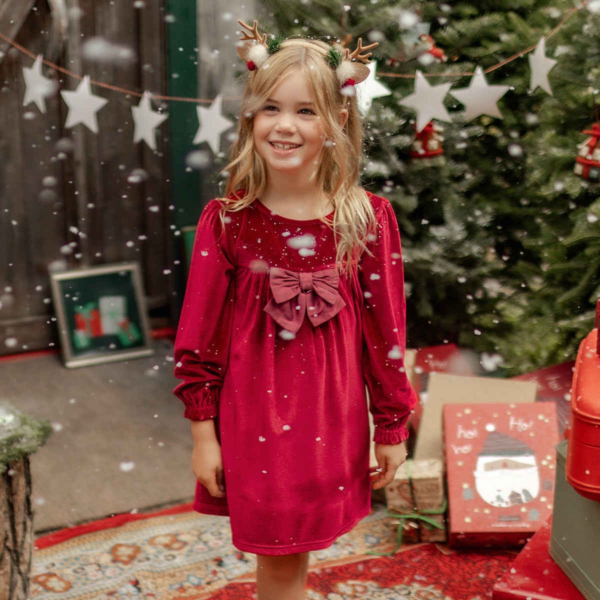 Fillette souriante portant une robe rouge en velours avec un nœud à l’avant, collection des Fêtes Souris Mini, entourée de décorations de Noël.