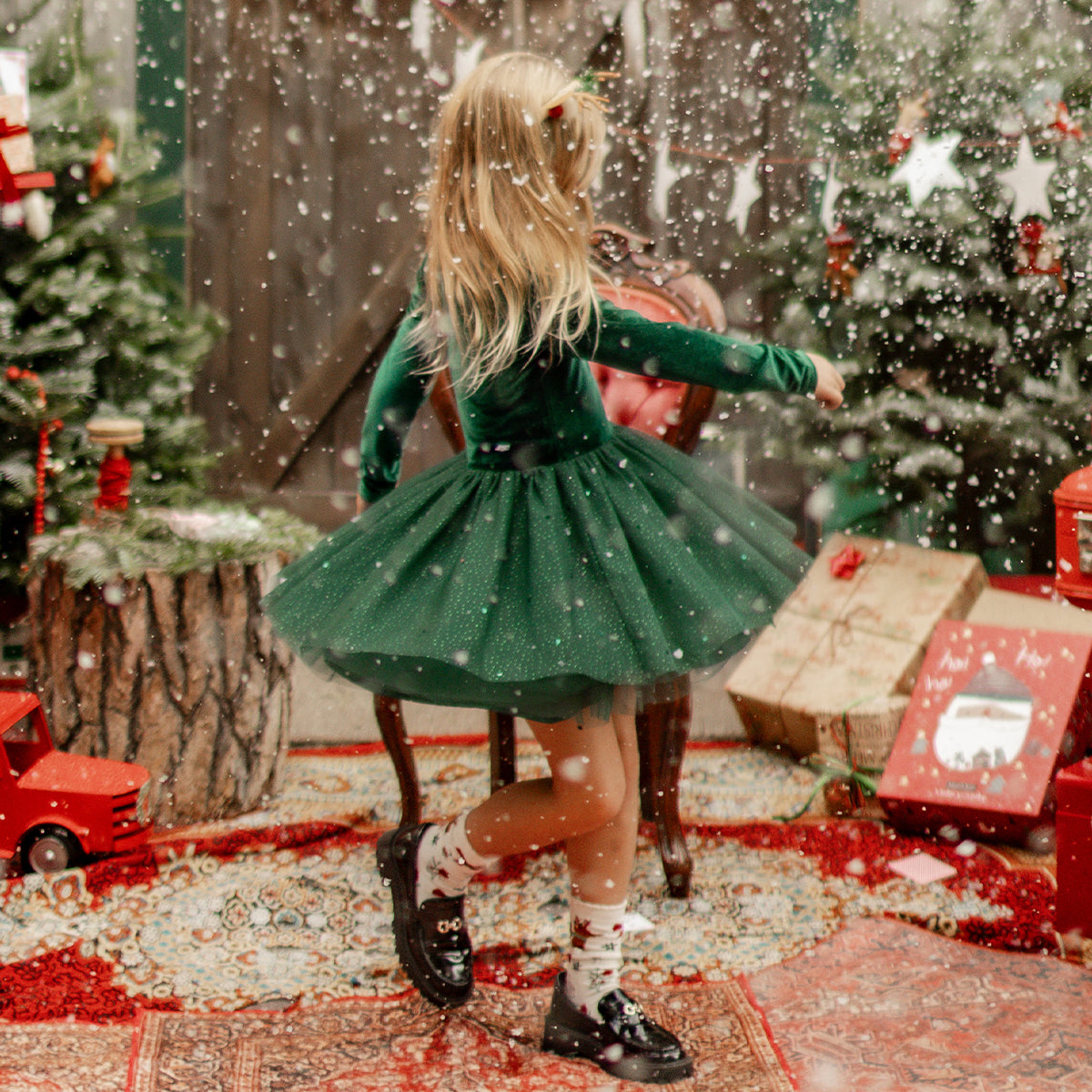 Petite fille portant une robe verte en velours et paillettes qui tourne joyeusement sous la neige, entourée de décorations de Noël scintillantes.