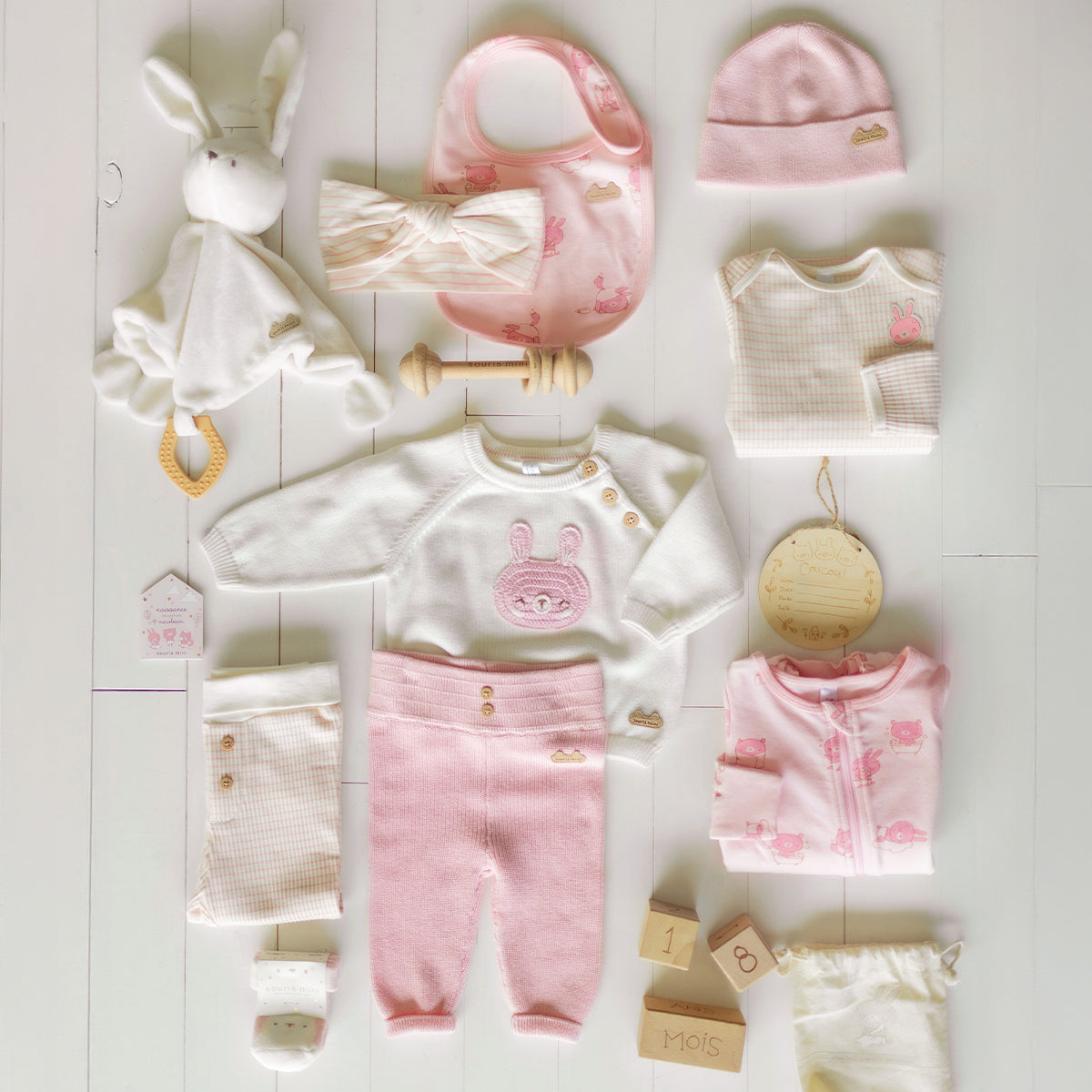 Ensemble cadeau pour bébé fille comprenant un pull blanc avec motif lapin rose, pantalon rose, body à carreaux crème et rose, grenouillère rose imprimée, bonnet rose, bavoir rose, bandeau à nouer, chaussettes, doudou lapin avec anneau de dentition, hochet en bois et accessoires de suivi de croissance sur fond blanc.
