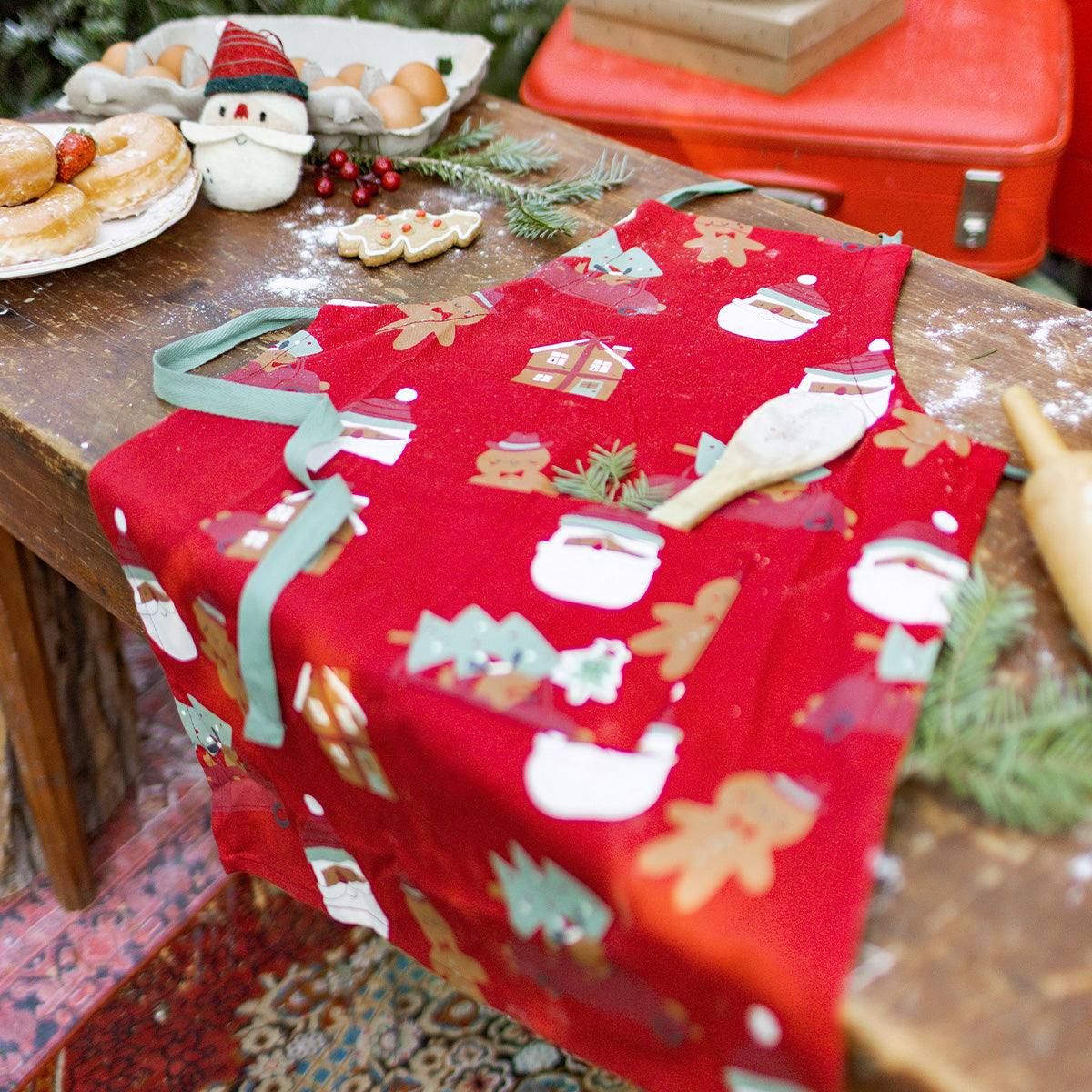 Tablier rouge à motifs festifs de Noël déposé sur une table de cuisine, entouré de biscuits et décorations, parfait pour cuisiner en famille.