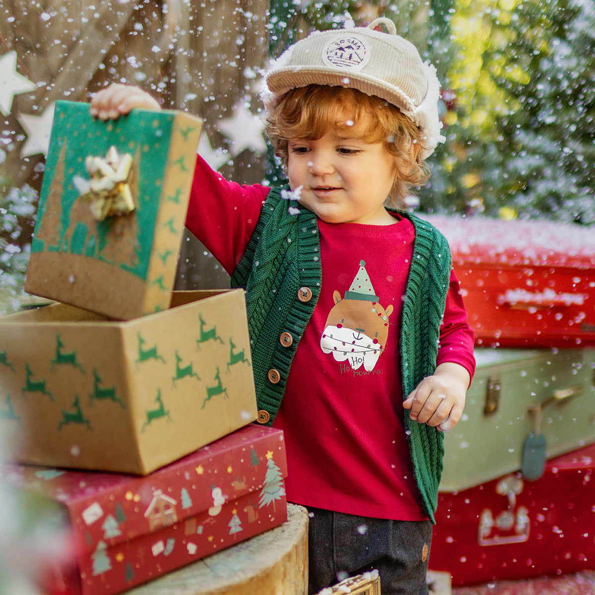 Petit garçon portant un t-shirt rouge avec imprimé d’ours de Noël et un gilet vert tricoté, jouant avec des boîtes cadeaux sous la neige, collection Souris Mini.