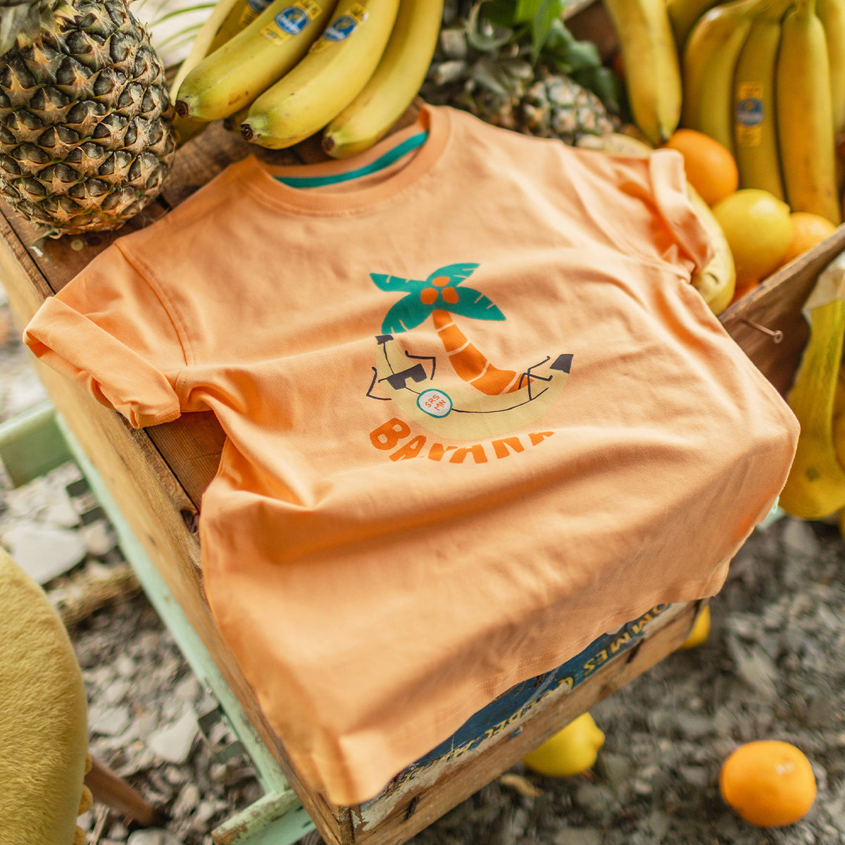 T-shirt orange avec illustration tropicale d’un palmier et d’une banane, posé sur une caisse en bois entourée de bananes, ananas et agrumes dans un décor estival. - Souris Mini