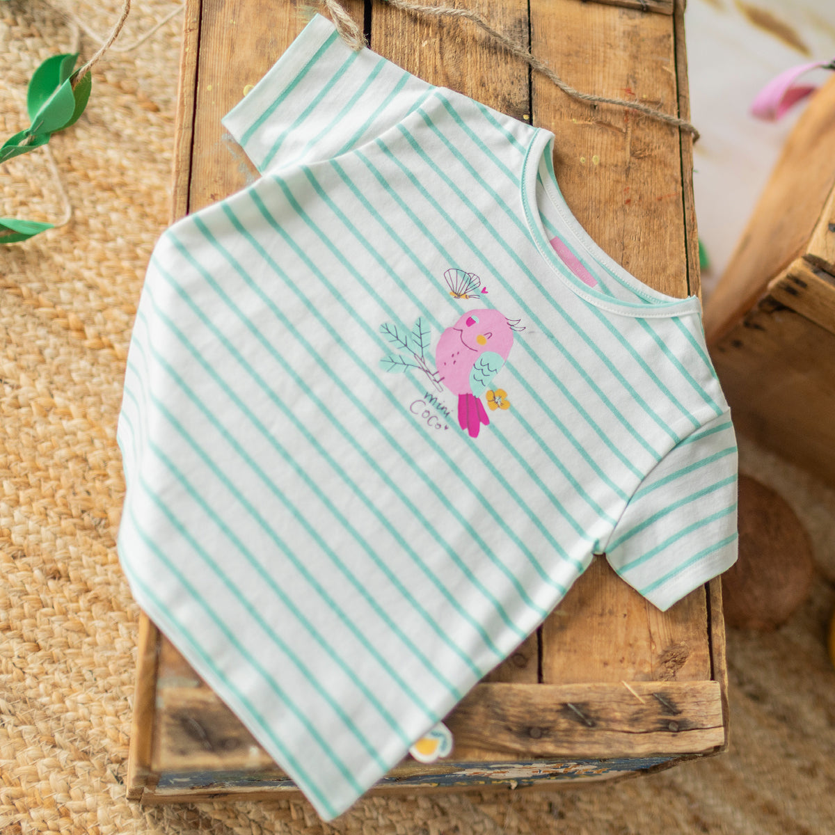 T-shirt rayé blanc et vert avec illustration d’un oiseau rose, présenté sur une caisse en bois dans un décor estival et tropical. - Souris Mini