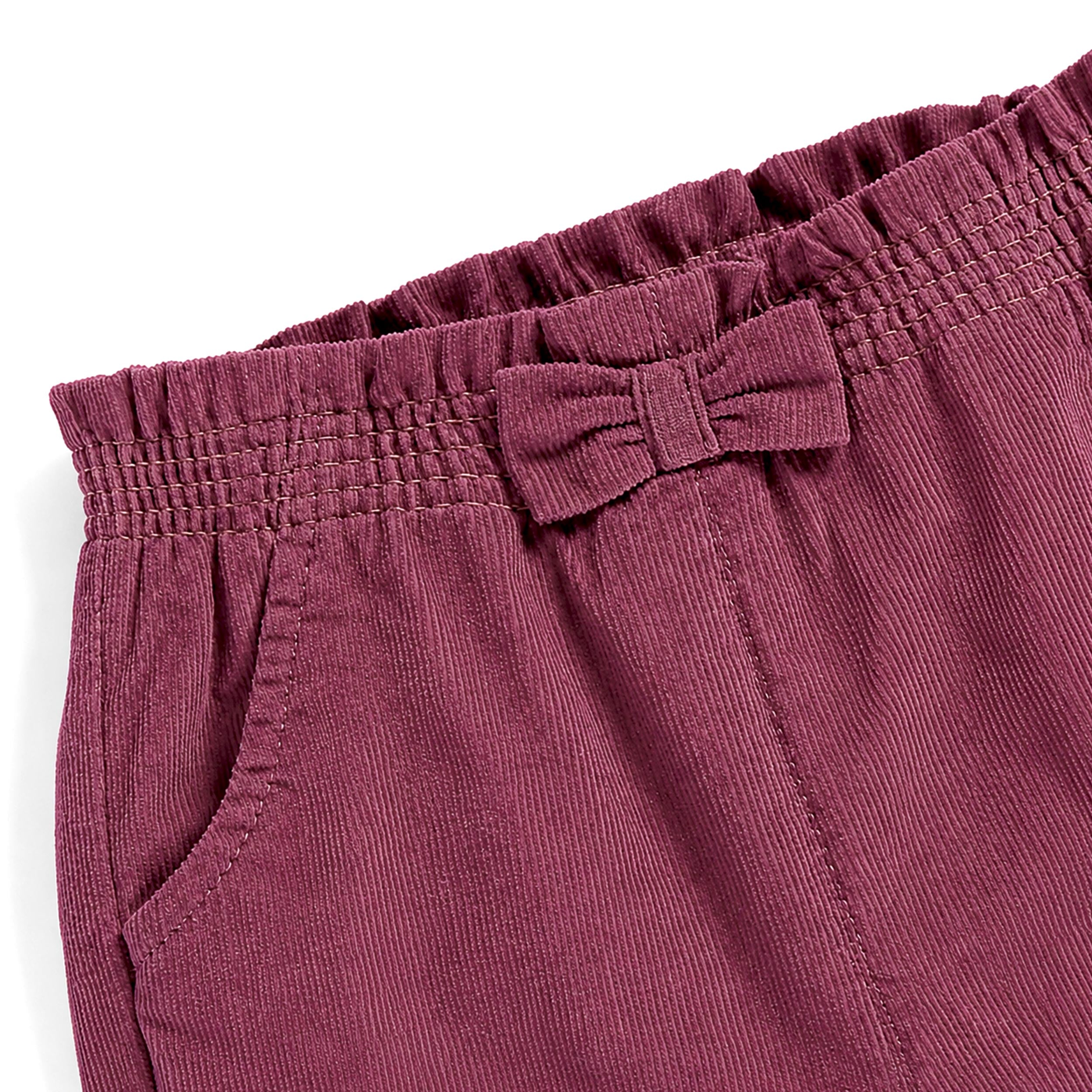 Gros plan sur la taille élastiquée à volants et le nœud décoratif d’un pantalon bébé fille en velours côtelé prune.