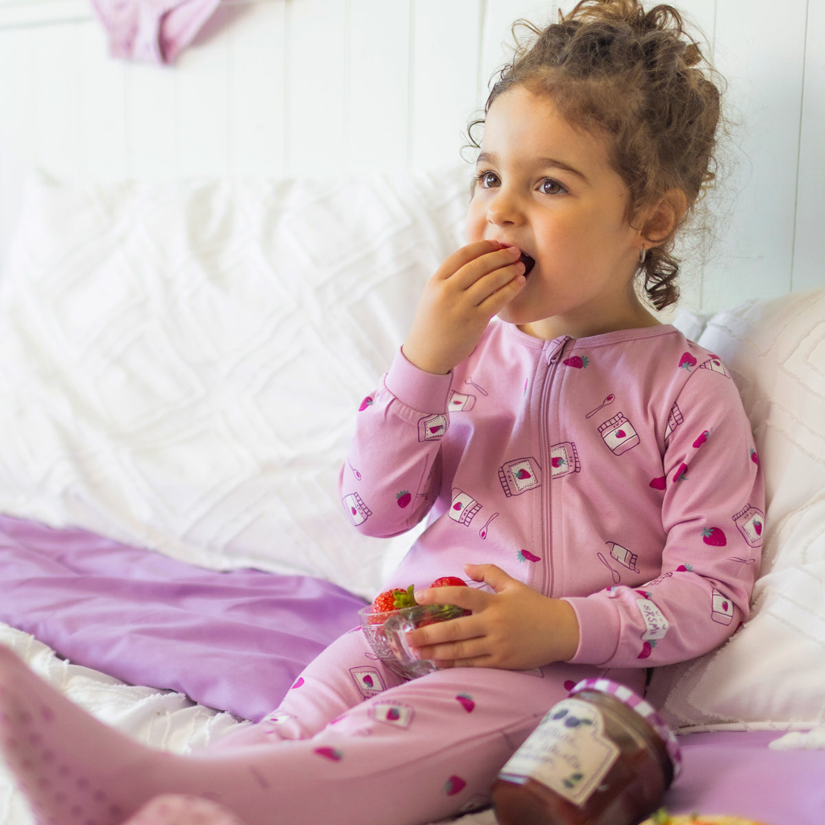 Pyjama une pièce mauve à petits motifs pour bébé en coton doux, porté par un bébé assis sur un lit, avec fermeture éclair pleine longueur et imprimé de fraises et pots de confiture.