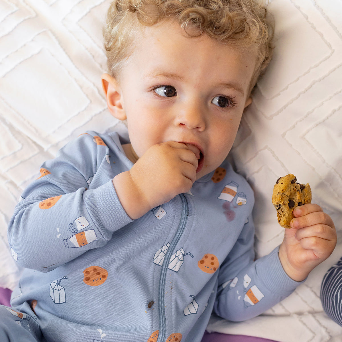 Bébé portant le pyjama une pièce bleu à petits motifs en coton doux, avec fermeture éclair et imprimés de biscuits et briques de lait, allongé sur un lit.