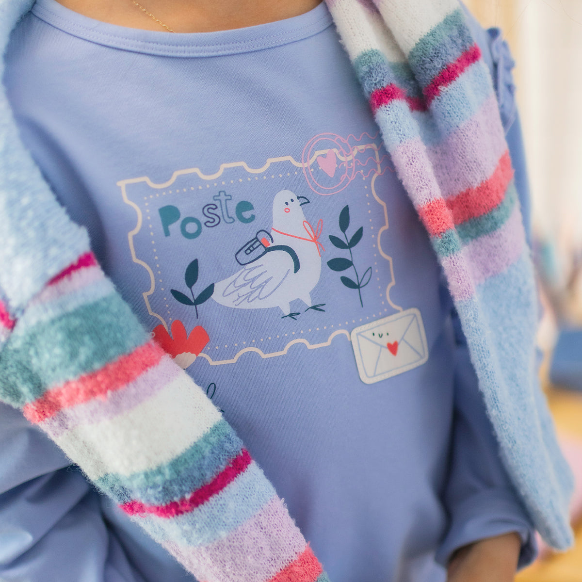 Gros plan d'un t-shirt bleu pour enfant avec illustration d'une colombe portant une lettre, inspirée d'un timbre-poste, porté sous un chandail de maille à rayures multicolores.
