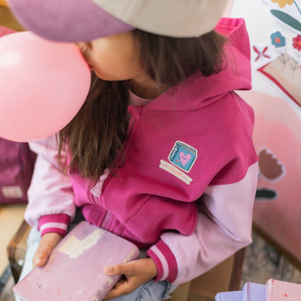 Veste rose en coton ouaté pour enfant avec capuchon, fermeture à glissière et écusson brodé sur la poitrine, portée par une enfant.