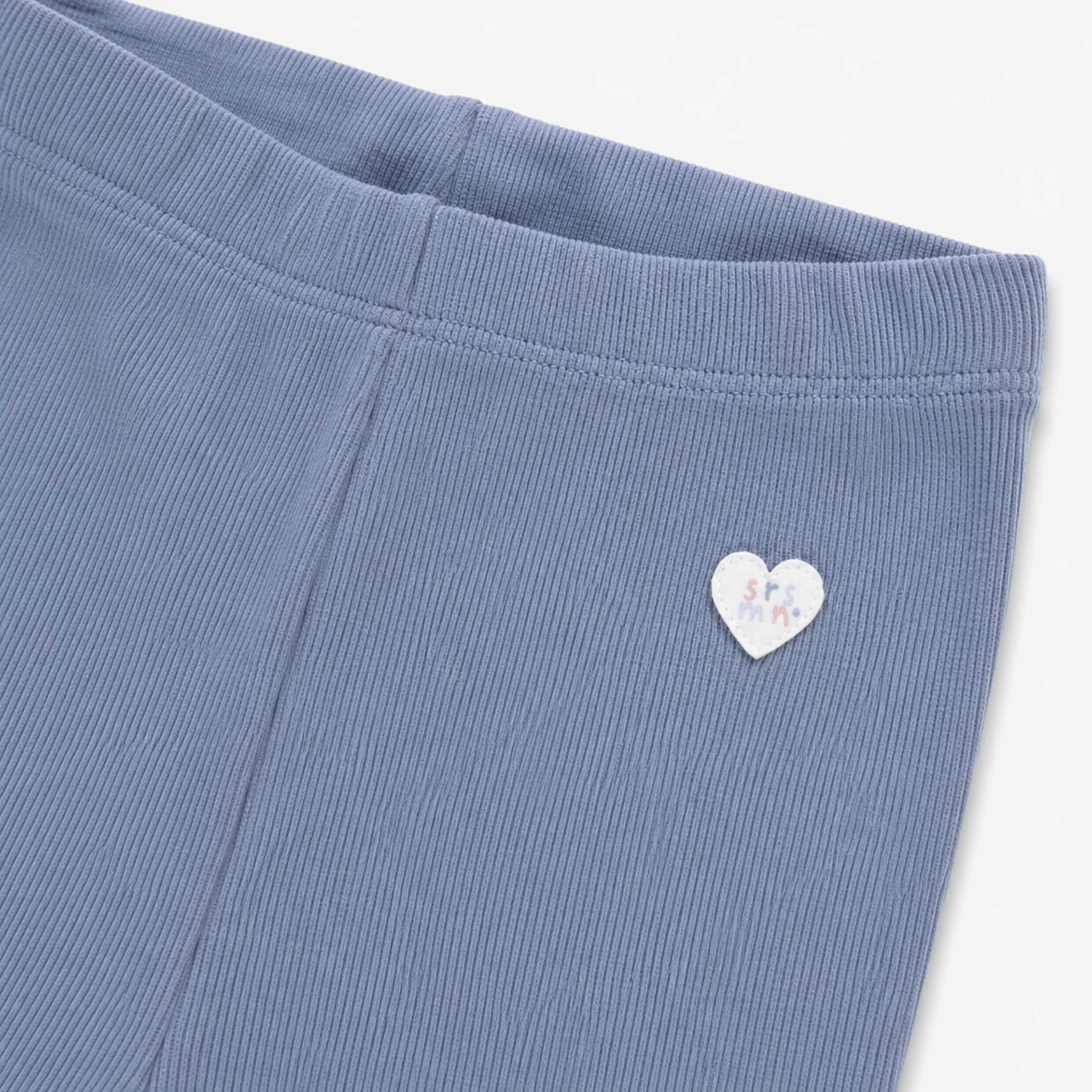 Gros plan sur un legging côtelé bleu pâle pour bébé fille, montrant la taille élastique et un petit écusson en forme de cœur cousu à la hanche.