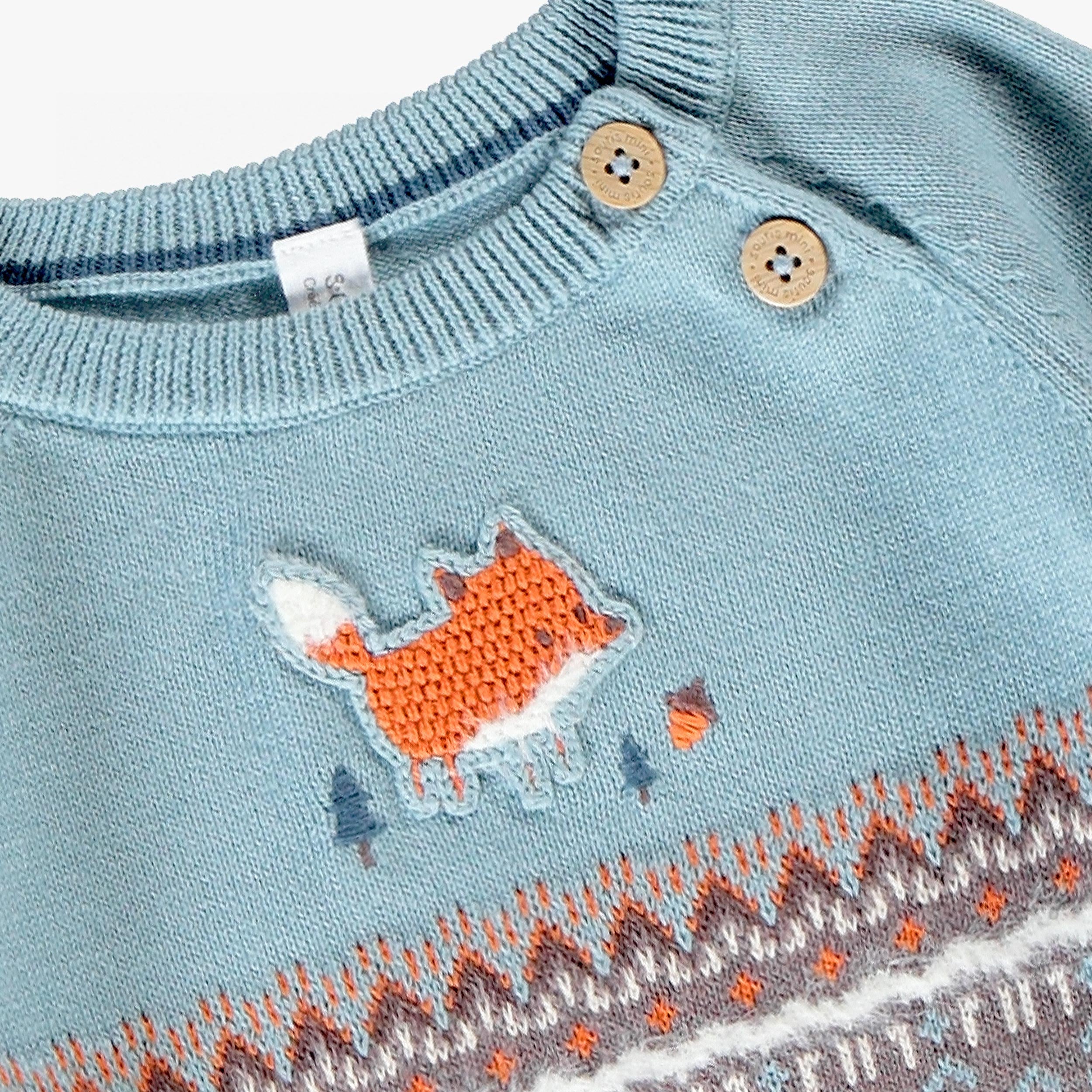 Gros plan sur un pull en tricot bleu et gris pour bébé garçon, mettant en valeur le motif de renard brodé, les boutons à l'épaule et les détails jacquard.