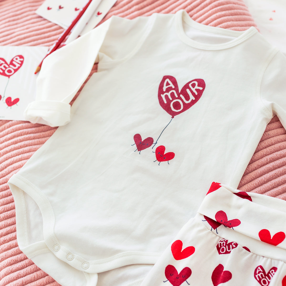 Cache-couche blanc pour bébé à motifs de cœurs rouges Souris Mini, avec pantalon assorti, présenté à plat sur un tissu rose.