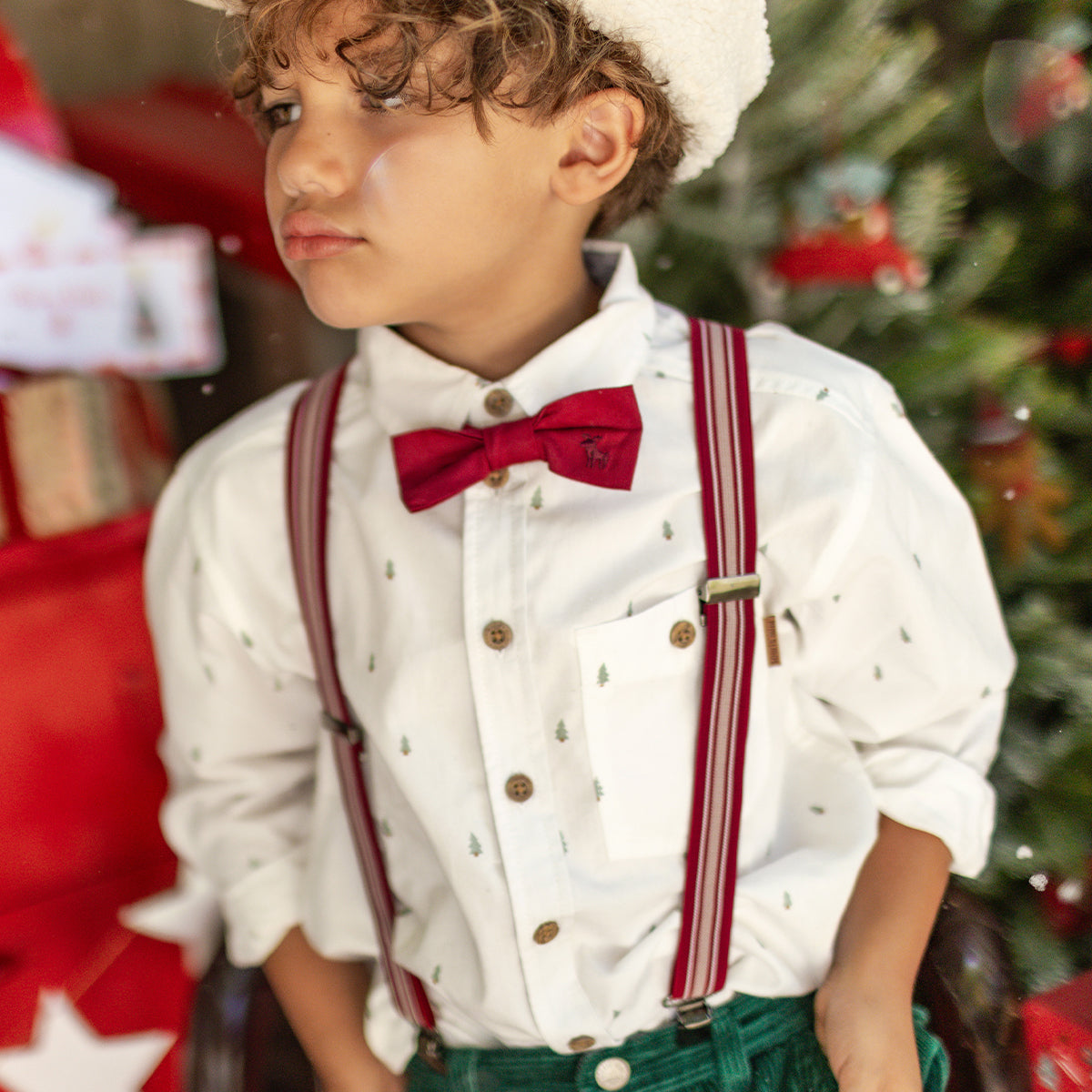Garçon portant une chemise blanche à motifs de sapins, bretelles rouges et nœud papillon, dans un décor festif de Noël, collection Souris Mini.