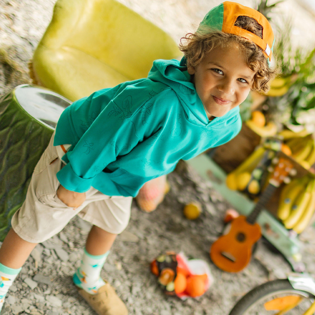 Enfant souriant portant un chandail à capuchon turquoise avec motifs tropicaux et une casquette orange et turquoise à l’envers, posant dans un décor de plage avec fruits et ukulélé.