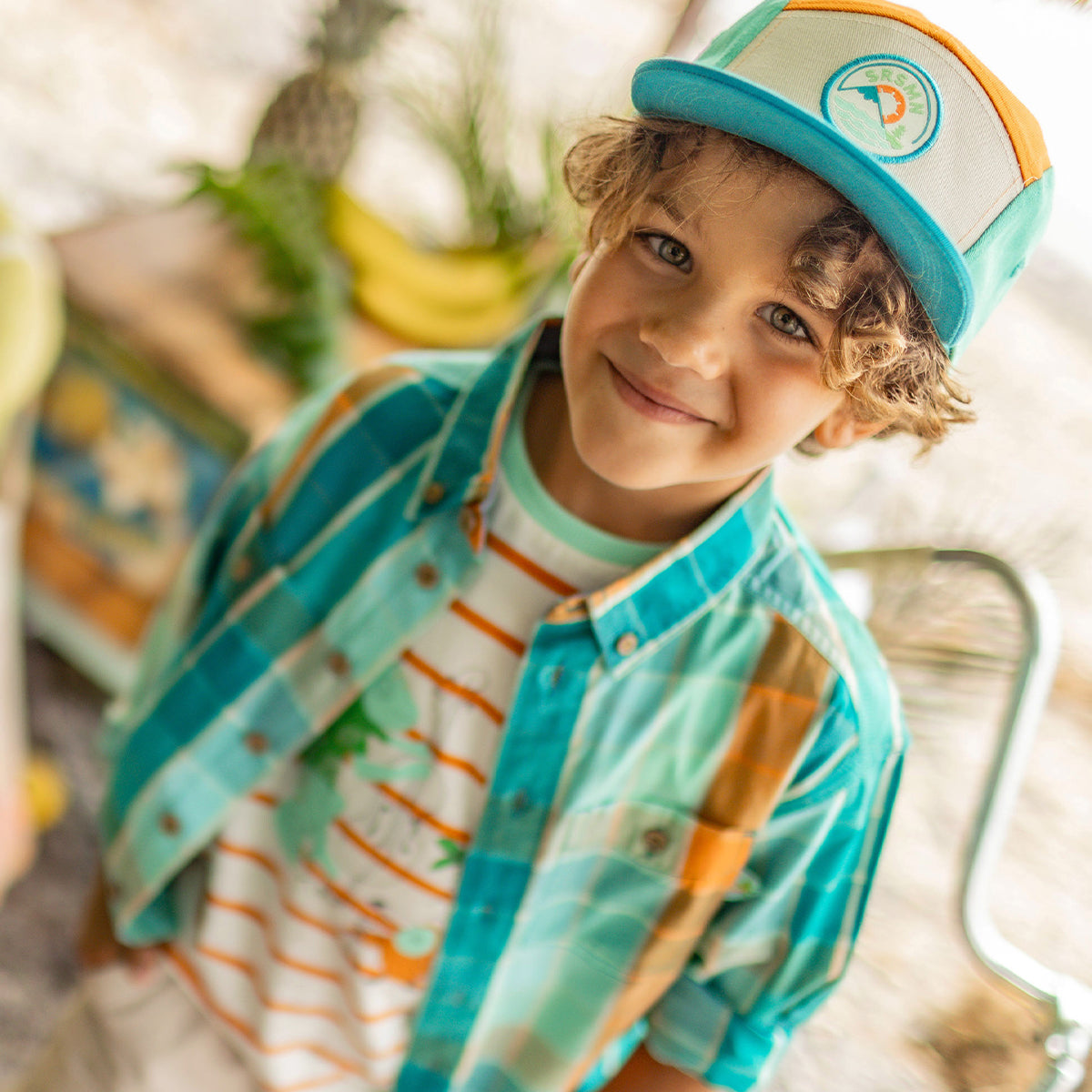 Enfant souriant en chemise turquoise-orange, t-shirt tropical et casquette bicolore, dans un décor de plage lumineux avec fruits exotiques. - Souris Mini