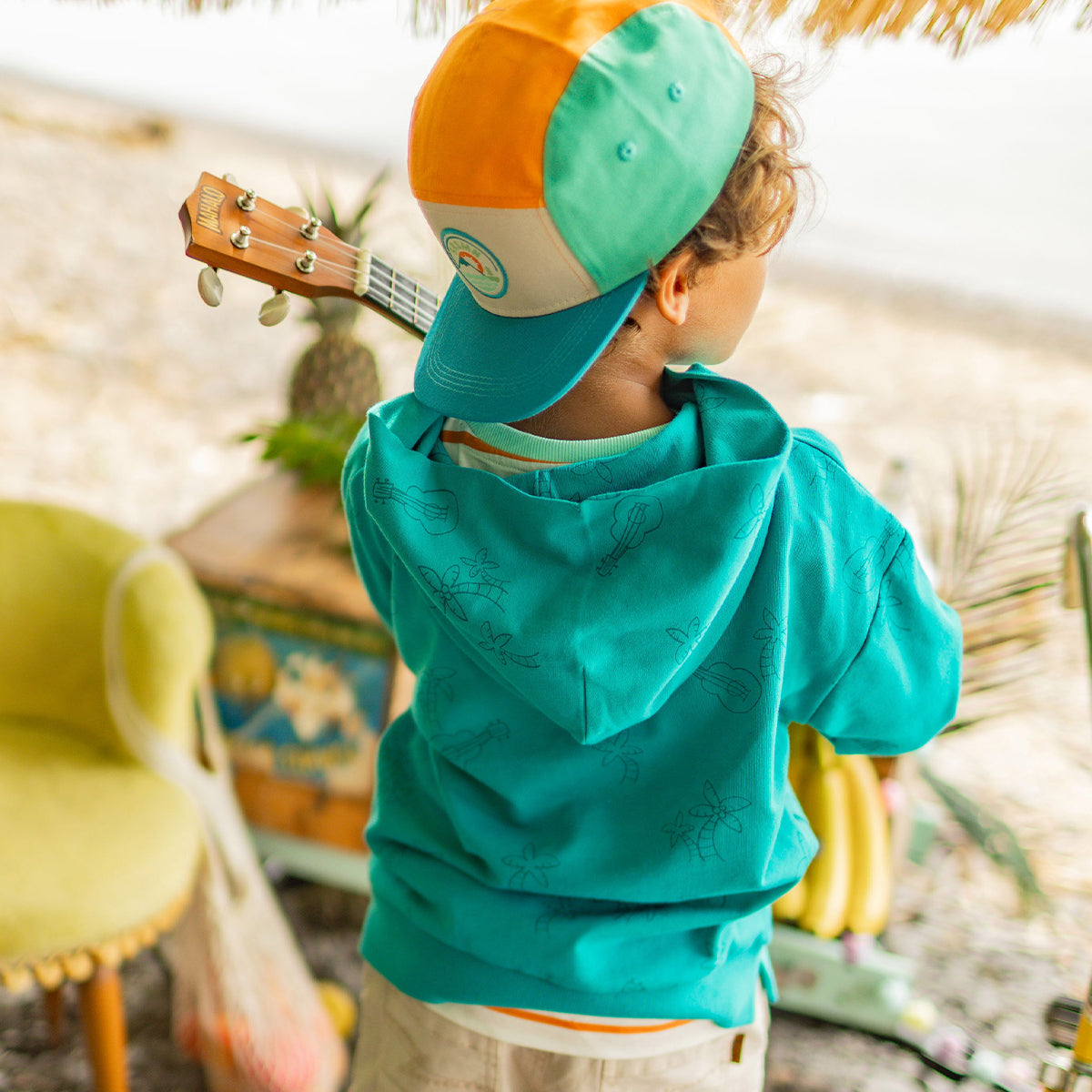Enfant vu de dos portant un chandail à capuchon turquoise avec motifs discrets et une casquette turquoise et orange, tenant un ukulélé dans un décor tropical près de la plage.