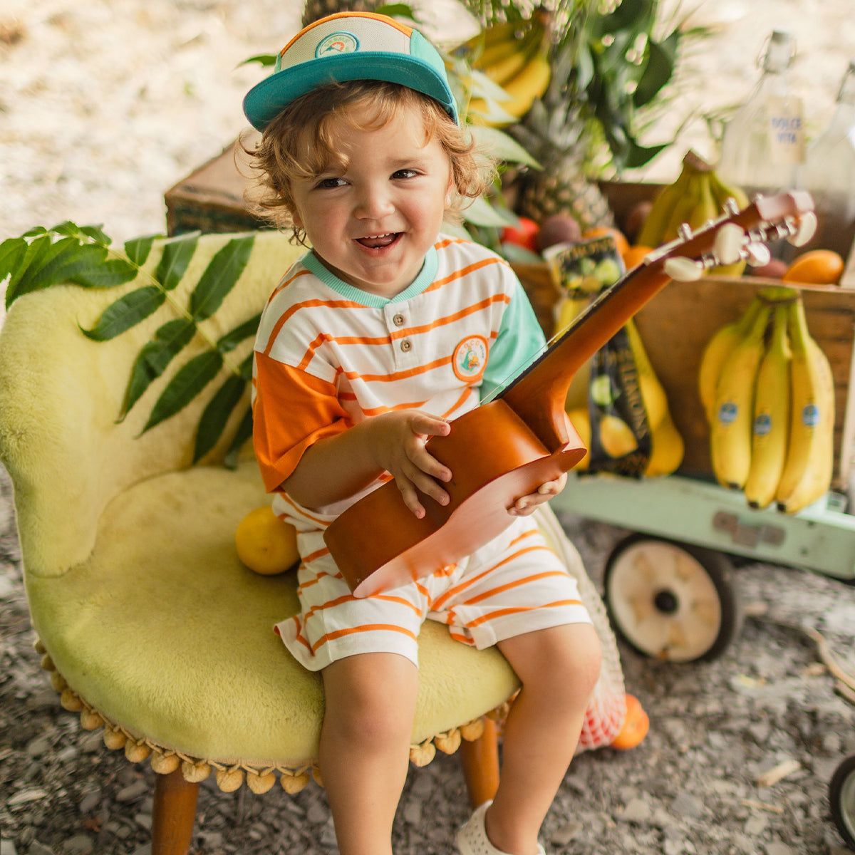 Jeune enfant souriant assis sur une chaise jaune, portant un ensemble rayé orange et turquoise, tenant un ukulélé dans un décor tropical avec des fruits.