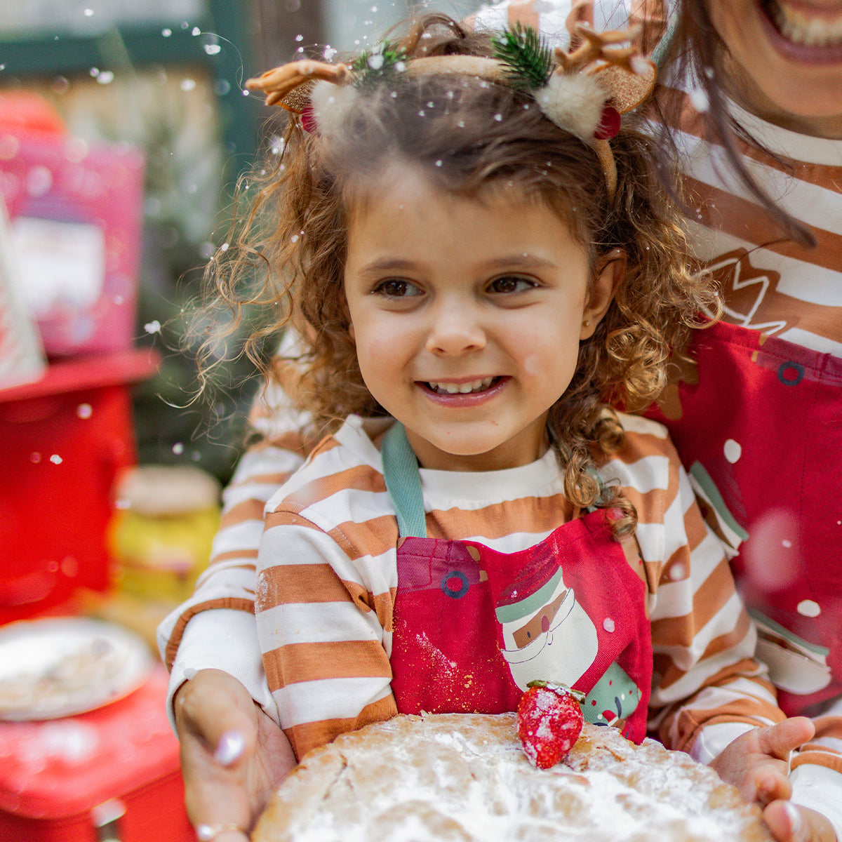 Fillette souriante portant un tablier rouge à motif de Père Noël et un pyjama rayé beige et blanc, tenant une tarte saupoudrée de sucre, ambiance pâtisserie de Noël, collection Souris Mini.