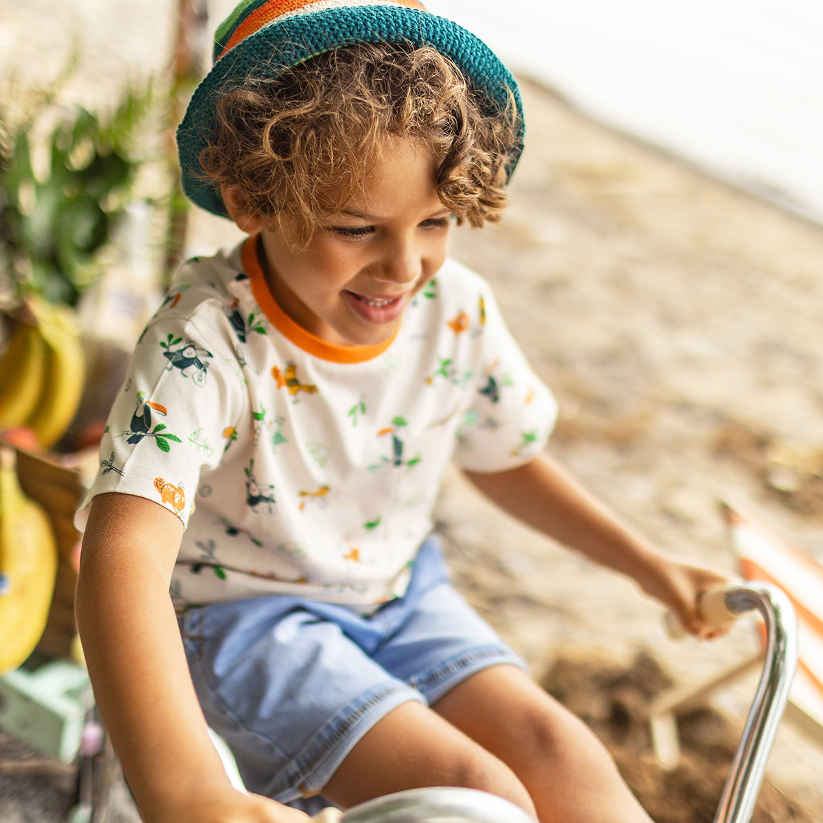 Enfant souriant portant un chapeau en crochet rayé turquoise-orange et un t-shirt crème à motifs tropicaux, assis sur un vélo près de la plage. - Souris Mini