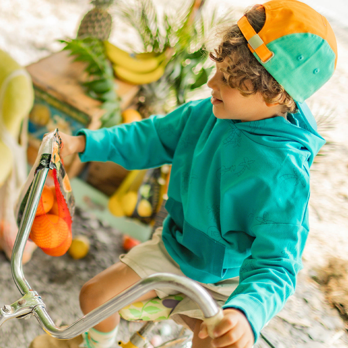 Enfant sur un vélo jaune portant un chandail à capuchon turquoise à motifs tropicaux et une casquette orange et turquoise à l’envers, avec un filet de fruits suspendu au guidon. - Souris Mini