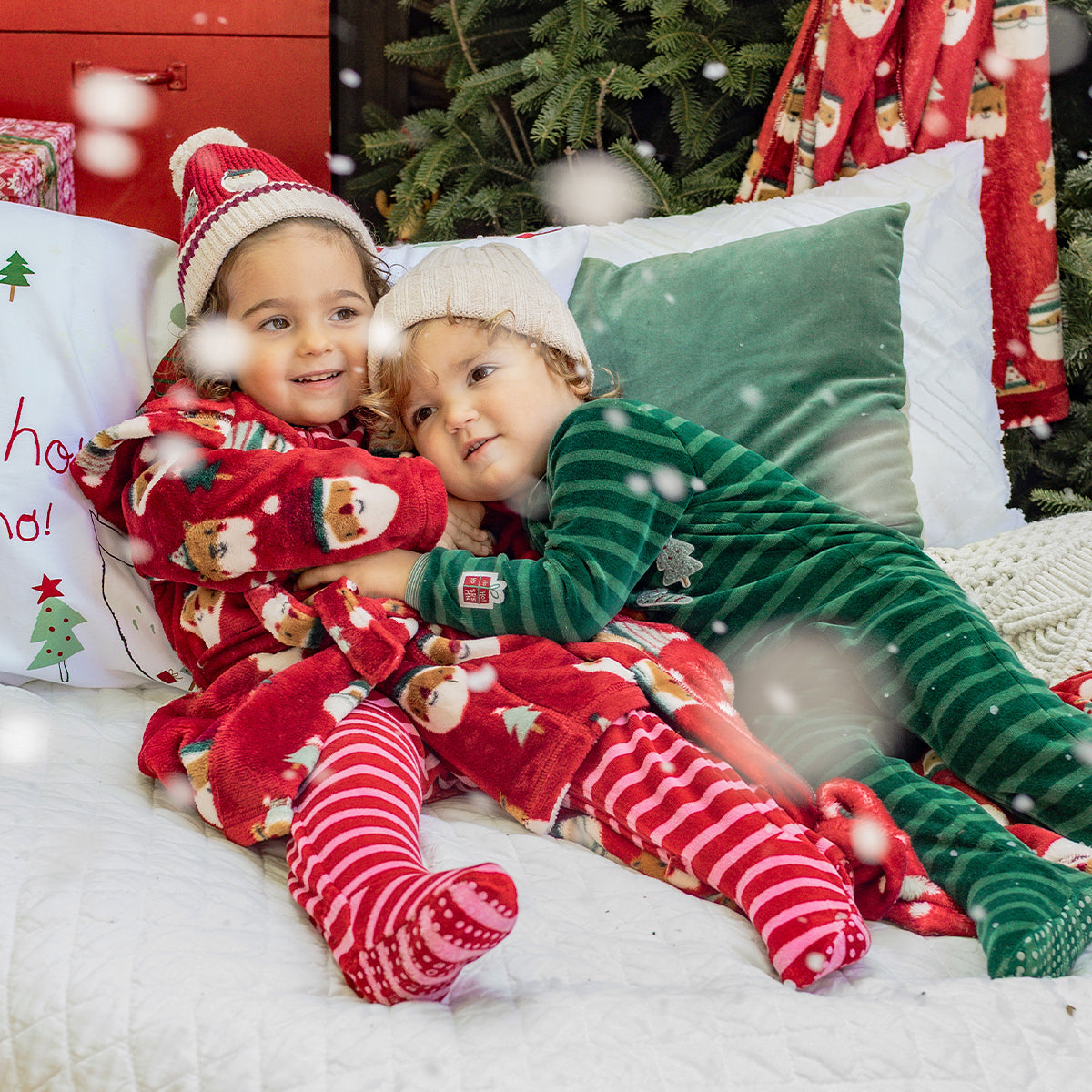 Deux enfants en pyjamas et robes de chambre festifs rouges et verts se blottissent ensemble sous la neige, dans une ambiance de Noël chaleureuse.