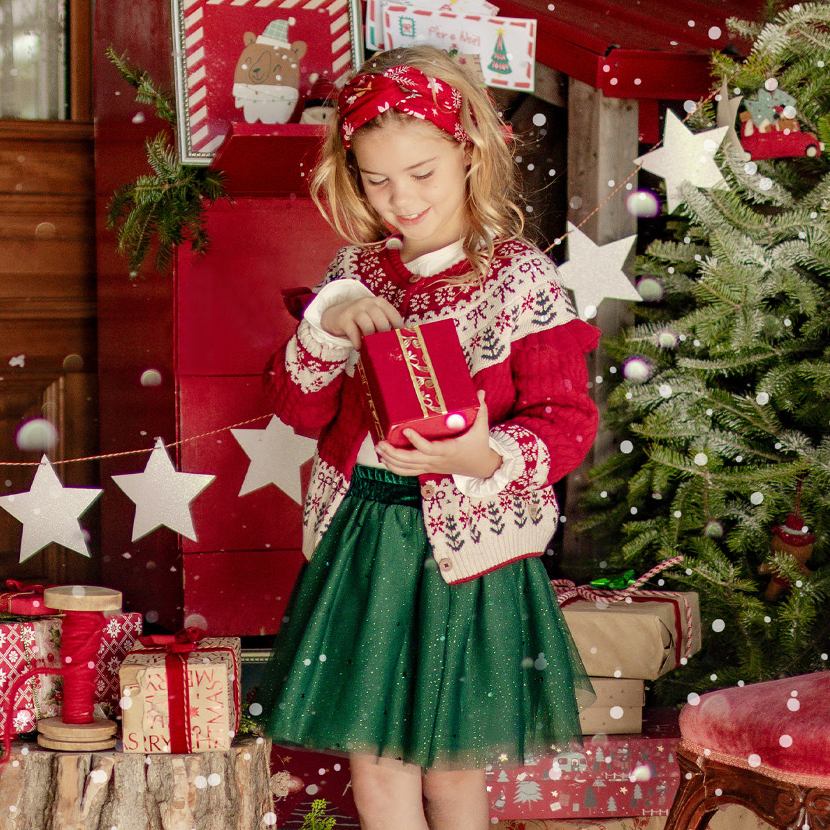 Petite fille vêtue d’un cardigan rouge à motifs festifs et d’une jupe verte scintillante ouvrant un cadeau devant un sapin de Noël décoré.
