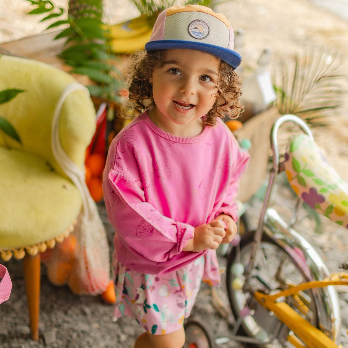 Fillette souriante portant un chandail rose et une jupe à motifs tropicaux, posant près d’un vélo décoré dans un décor estival.