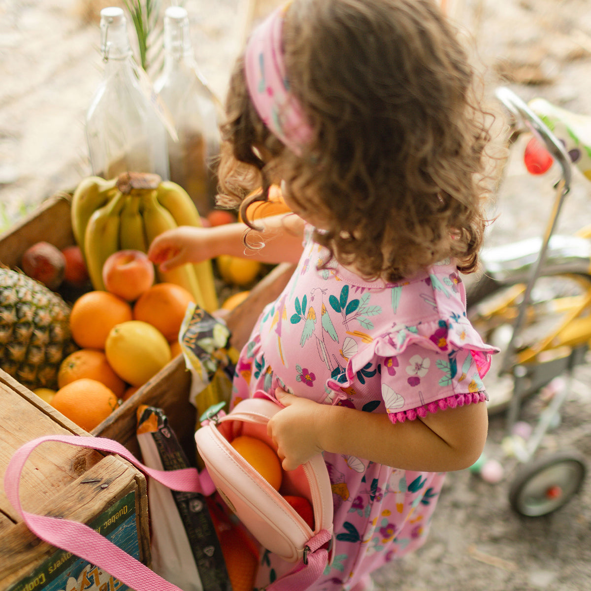 Fillette en robe rose à motifs tropicaux ramassant des fruits dans un étal coloré.