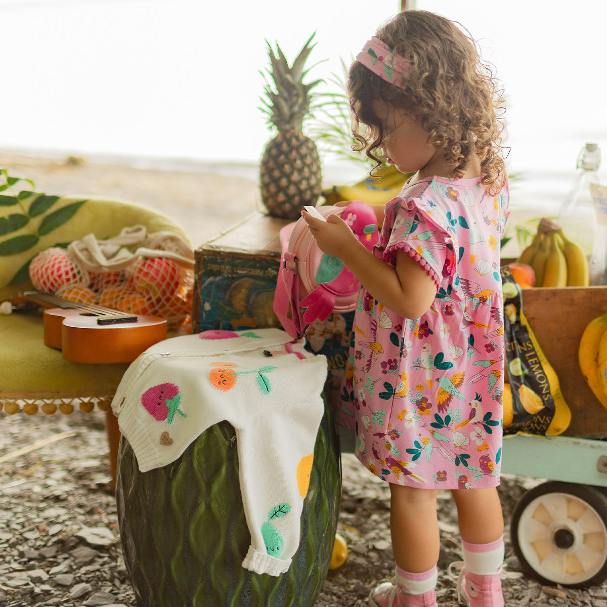 Fillette en robe rose à motifs tropicaux tenant un petit sac, avec un cardigan blanc à fruits posé devant elle dans un décor estival avec ananas et bananes.