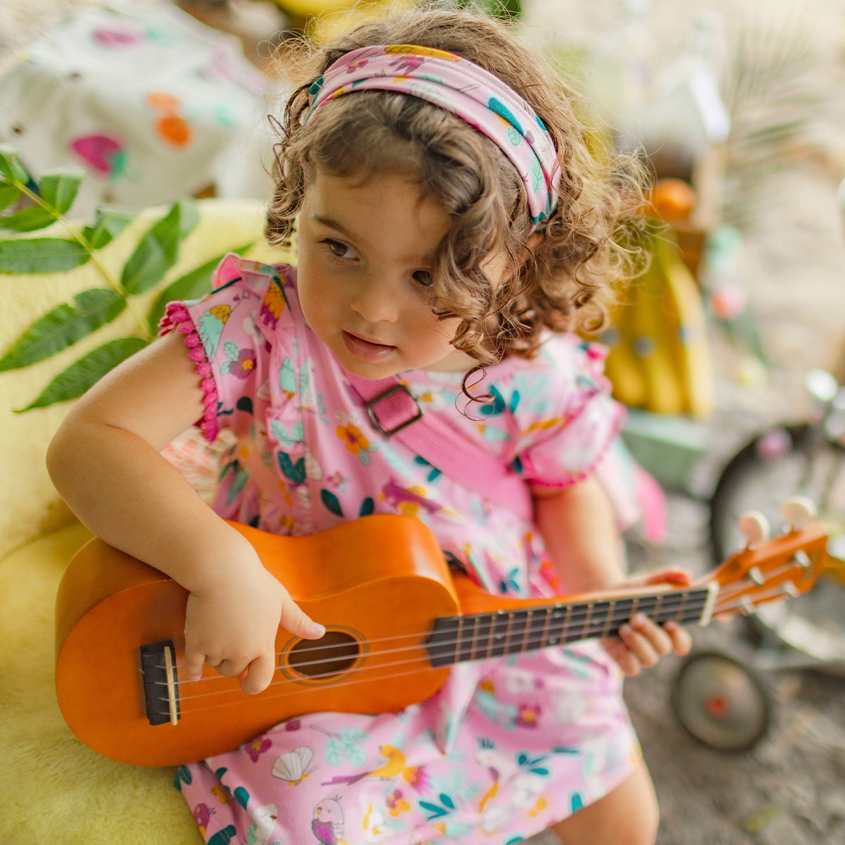 Fillette en robe rose à motifs tropicaux jouant du ukulélé, assise dans un décor estival coloré.
