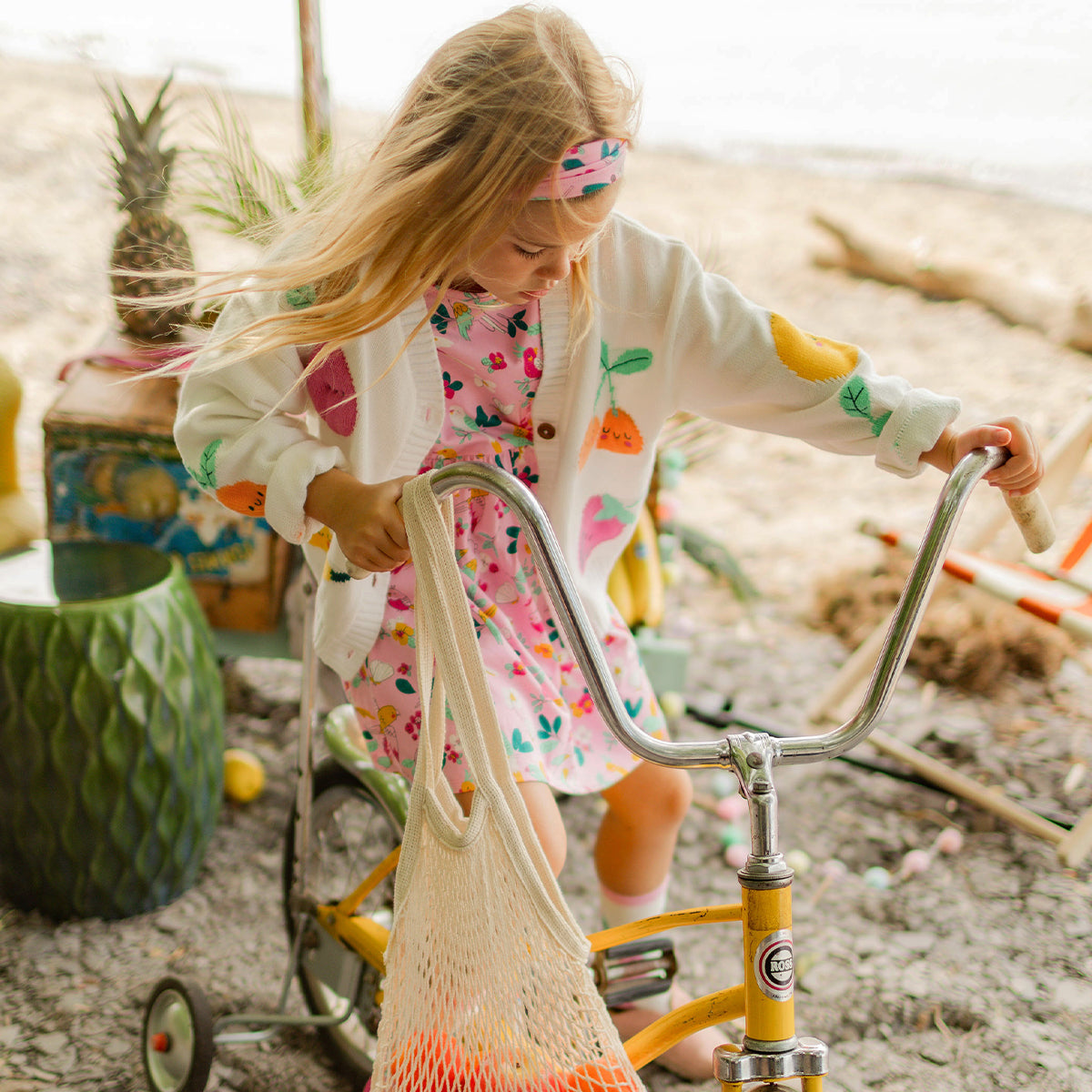 Fillette en robe rose à motifs fruités portant un cardigan crème décoré de fruits et un bandeau assorti, montant sur un vélo jaune avec un filet de fruits, dans un décor de plage. - Souris Mini