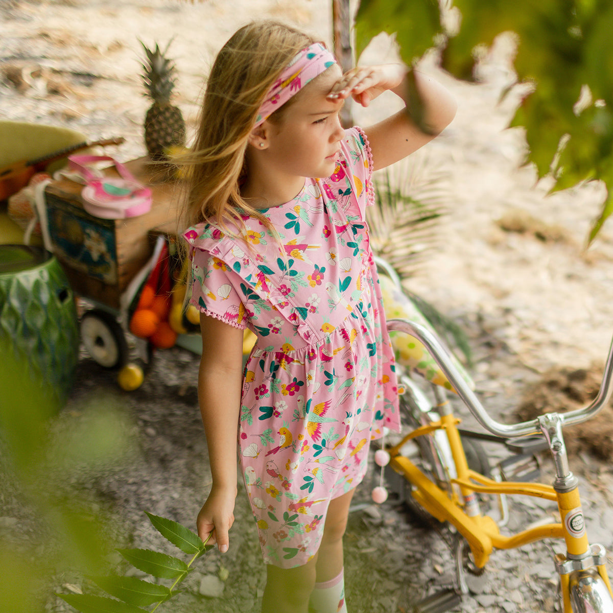 Fillette en robe rose à motifs tropicaux avec volants et bandeau assorti, regardant au loin près d’un vélo jaune dans un décor de plage entouré de végétation.