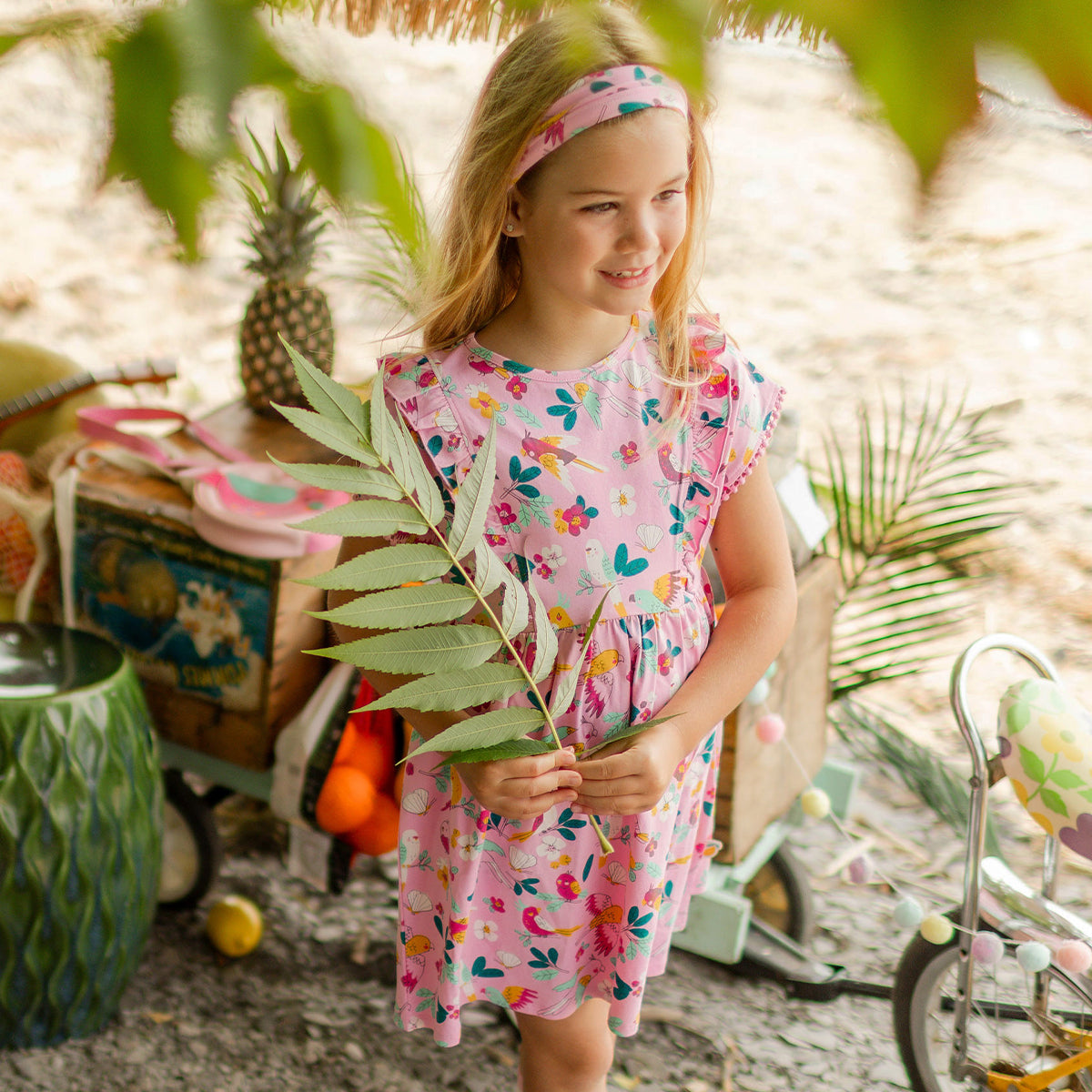 Fillette en robe rose à motifs tropicaux et bandeau assorti tenant une grande feuille, debout près d’un vélo jaune dans un décor lumineux inspiré de la plage. - Souris Mini
