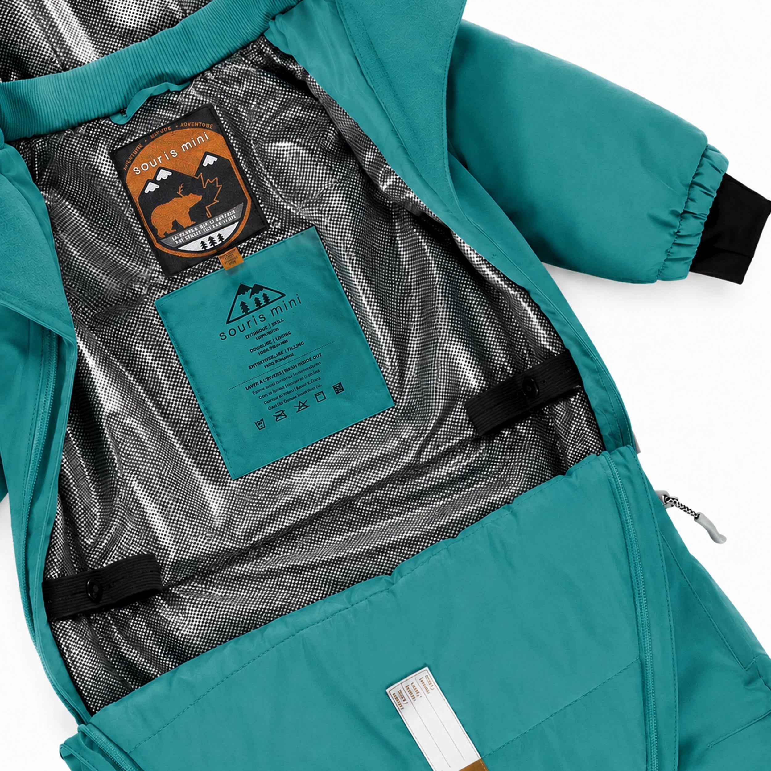 Gros plan sur l’intérieur d’une combinaison de neige bébé turquoise montrant la doublure thermoréfléchissante, les sangles de maintien et les finitions isolantes.