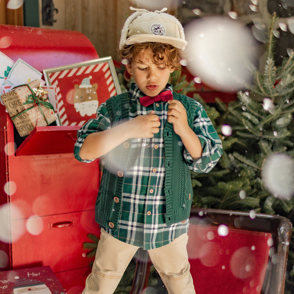 Petit garçon habillé pour Noël avec une chemise verte à carreaux, un débardeur en tricot, un nœud papillon rouge et une casquette, dans un décor festif.