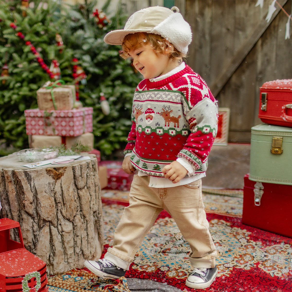 Petit garçon portant un pull rouge et gris à motifs de Noël avec rennes et Père Noël, marchant joyeusement dans un décor festif avec sapins et cadeaux, collection hiver Souris Mini.