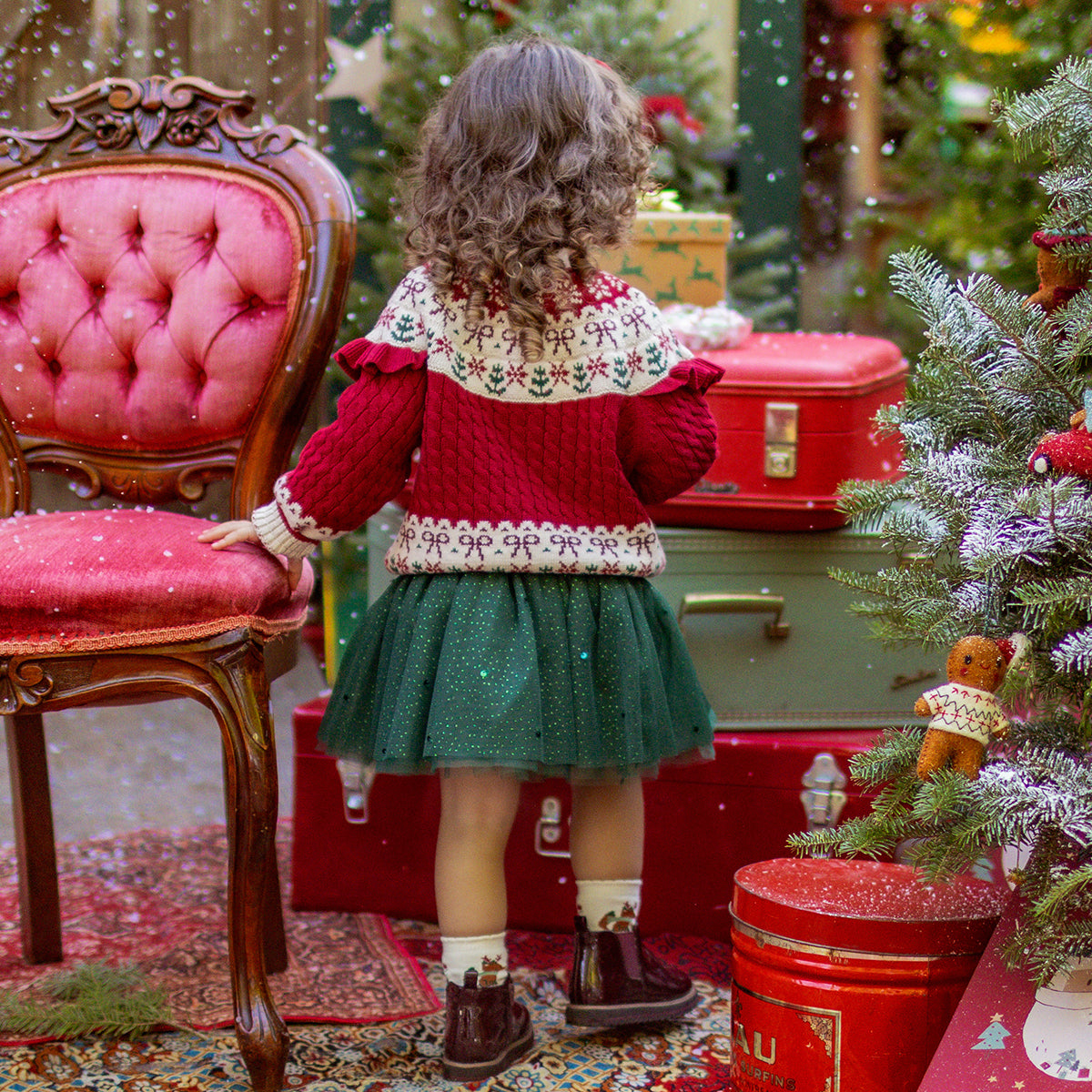 Petite fille vue de dos portant un pull tricoté rouge et blanc à motifs de Noël avec une jupe verte scintillante, dans un décor hivernal chaleureux, collection Souris Mini.