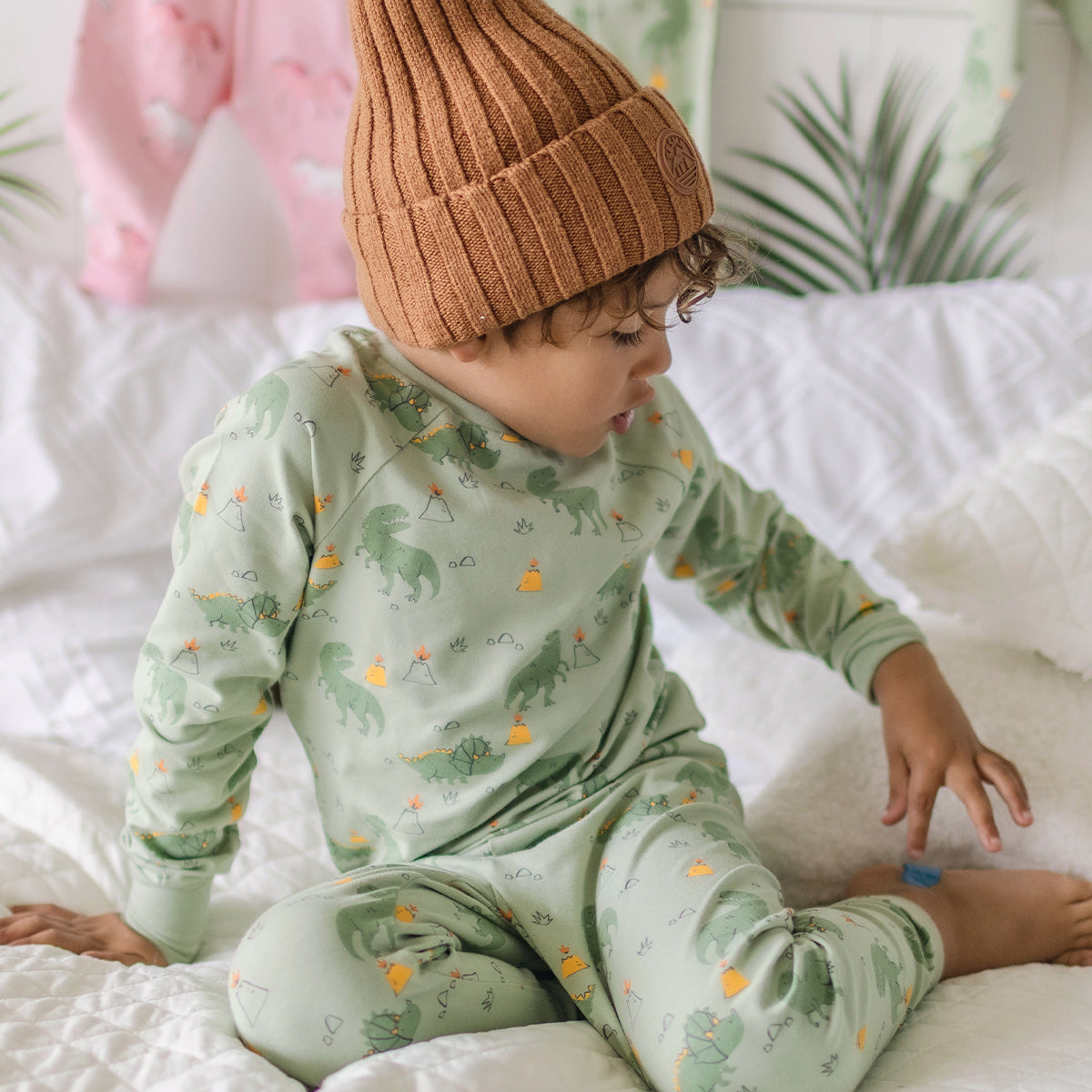 Garçon portant un pyjama vert à motifs de dinosaures Souris Mini, assis sur un lit blanc et jouant calmement.