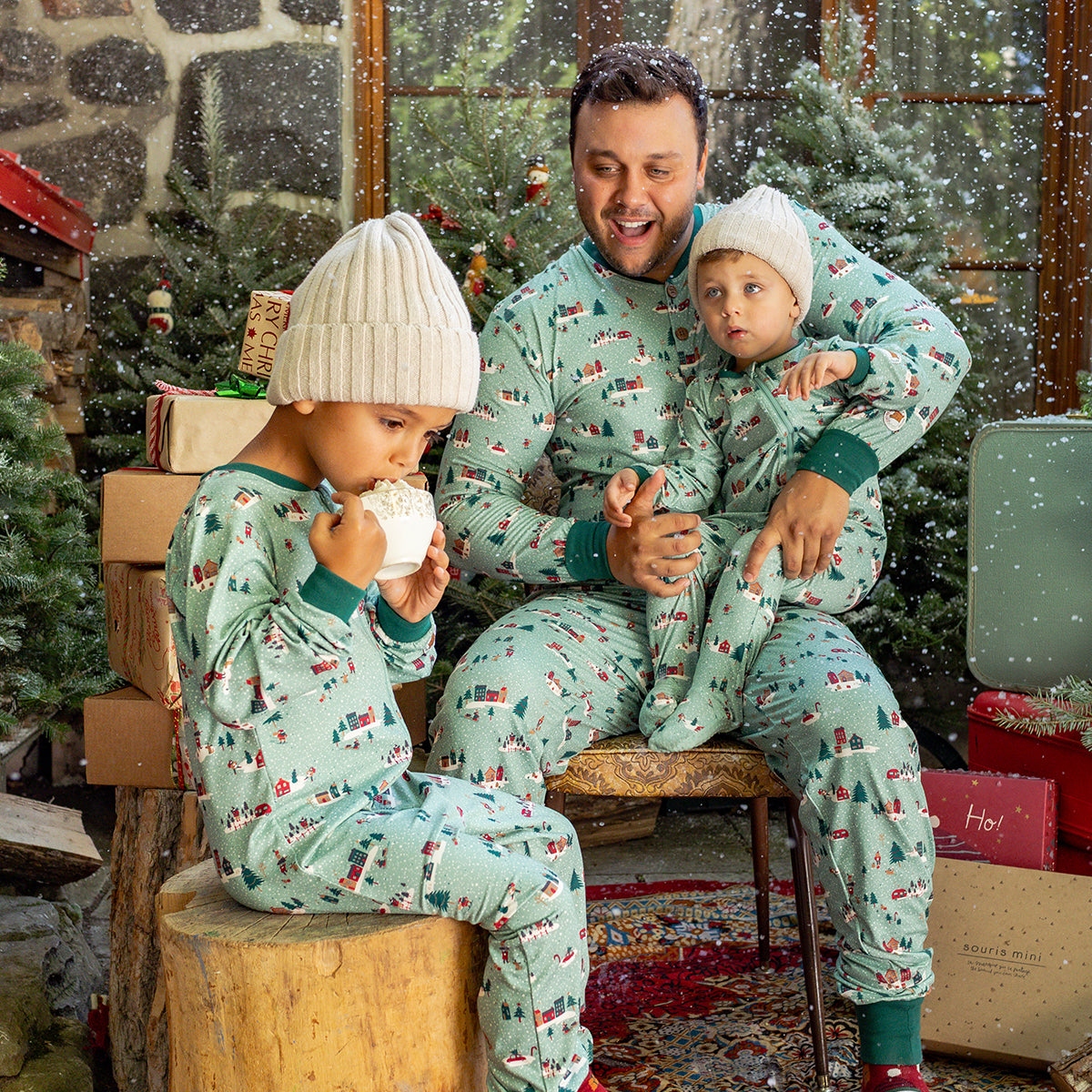 Famille portant des pyjamas verts à motifs des Fêtes, profitant d’un moment cocooning dans un décor hivernal avec sapins et cadeaux.