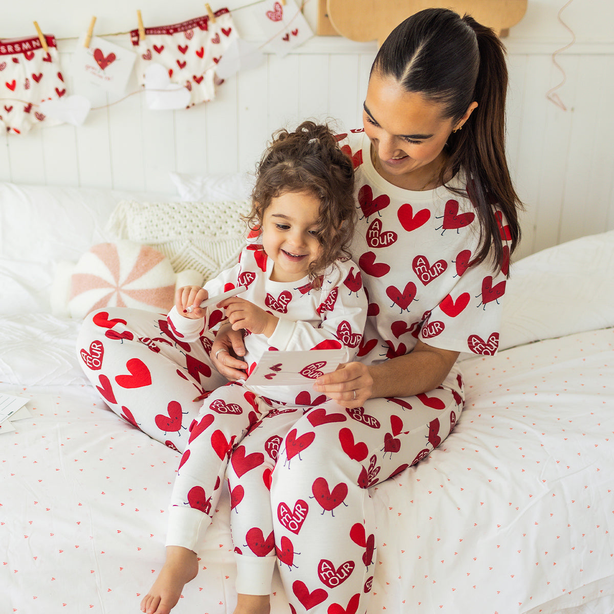 Maman et enfant portant des pyjamas assortis blancs à motifs de cœurs rouges Souris Mini, assis sur un lit et partageant un moment complice.