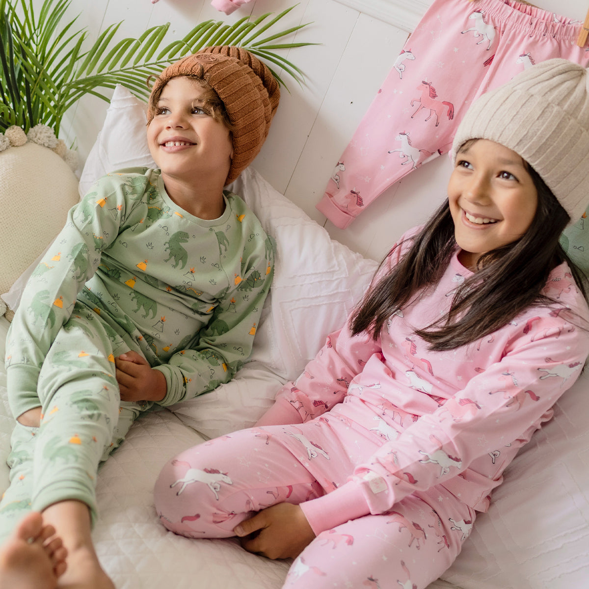 Deux enfants souriants portant des pyjamas Souris Mini, l’un vert à motifs de dinosaures et l’autre rose à motifs de licornes, assis sur un lit blanc.