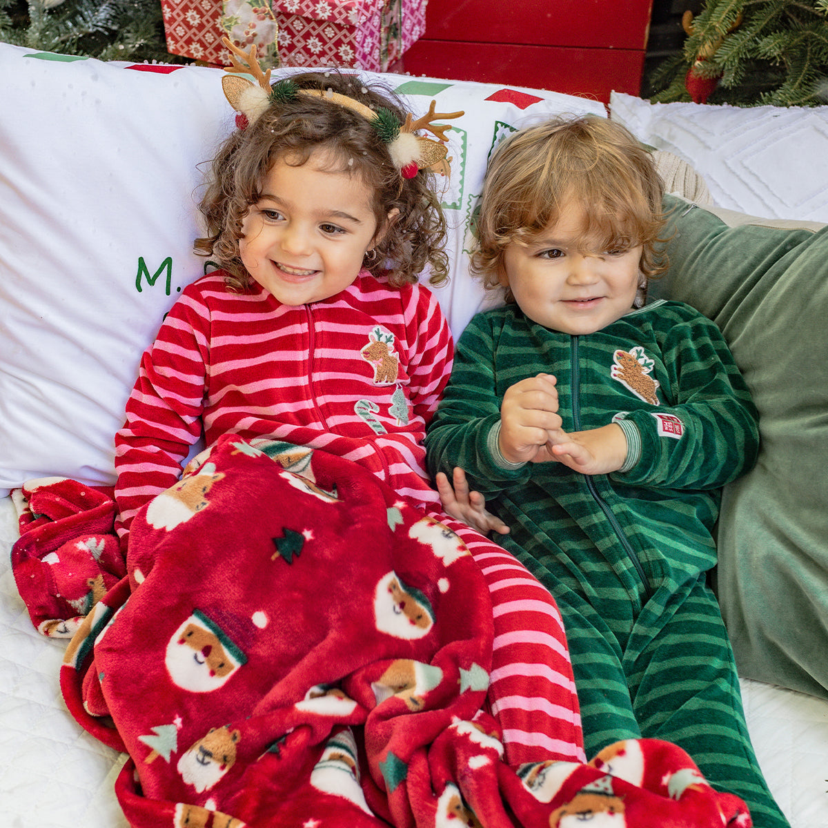 Deux enfants en pyjamas de Noël à rayures rouges et vertes grimpant sur un lit enneigé, entourés de couvertures festives et d’un sapin décoré.