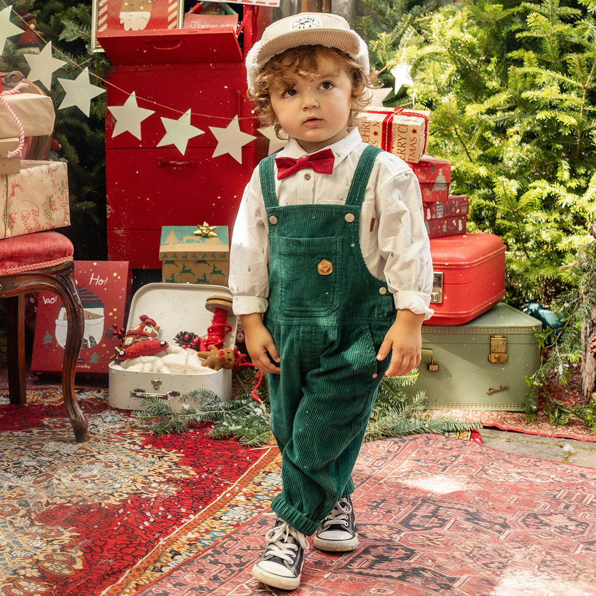 Petit garçon portant une salopette en velours verte, une chemise blanche et un nœud papillon rouge, dans un décor festif de Noël, collection Souris Mini.