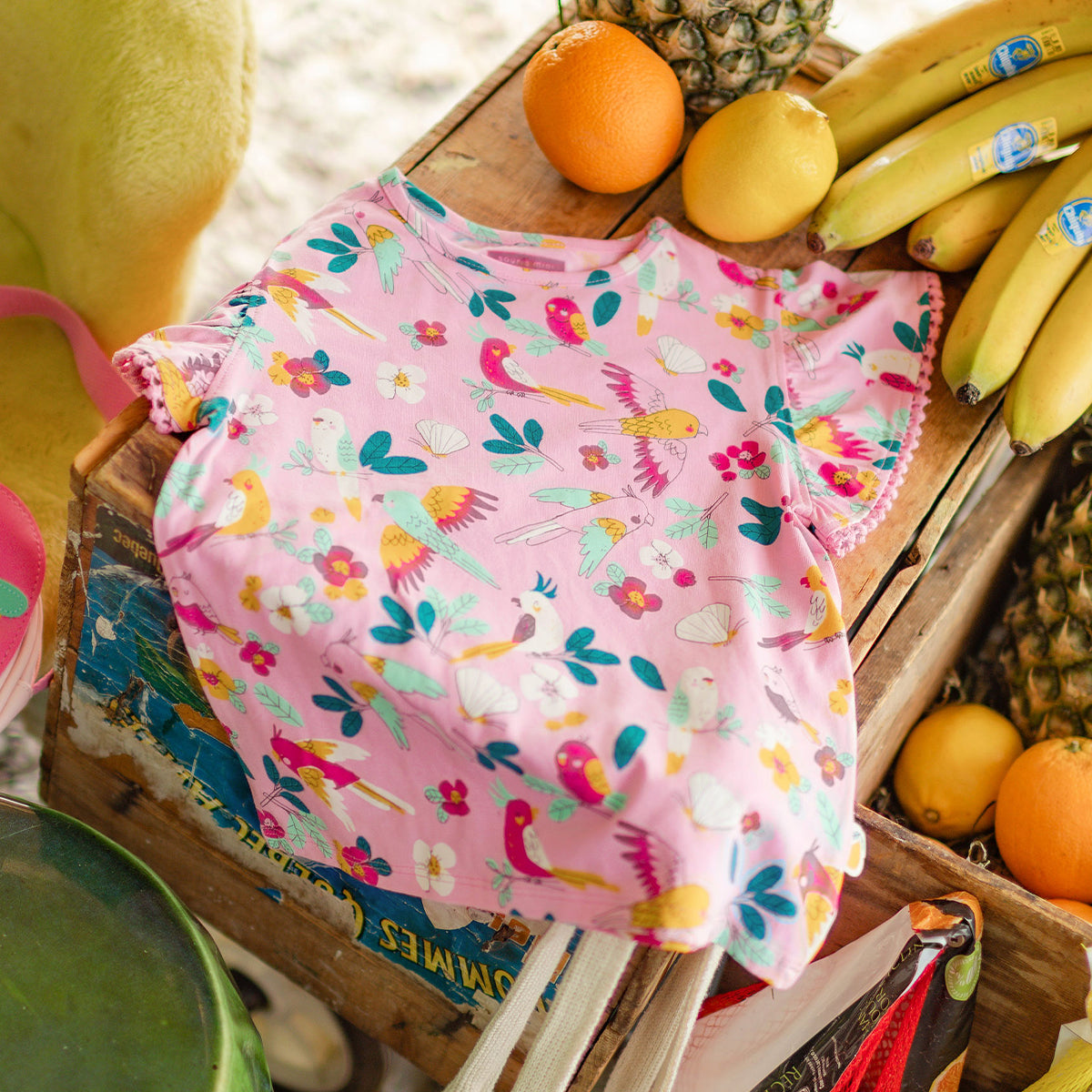 T-shirt rose pour fillette avec imprimé coloré d’oiseaux et de fleurs, présentée sur une caisse en bois avec des fruits tropicaux.