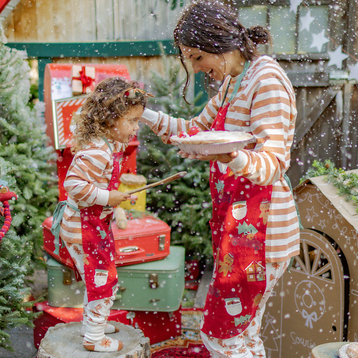 Maman et fille portant des tabliers rouges à motifs de Noël et des pyjamas rayés beige et blanc, partageant un moment de pâtisserie festif dans un décor hivernal, collection Souris Mini.