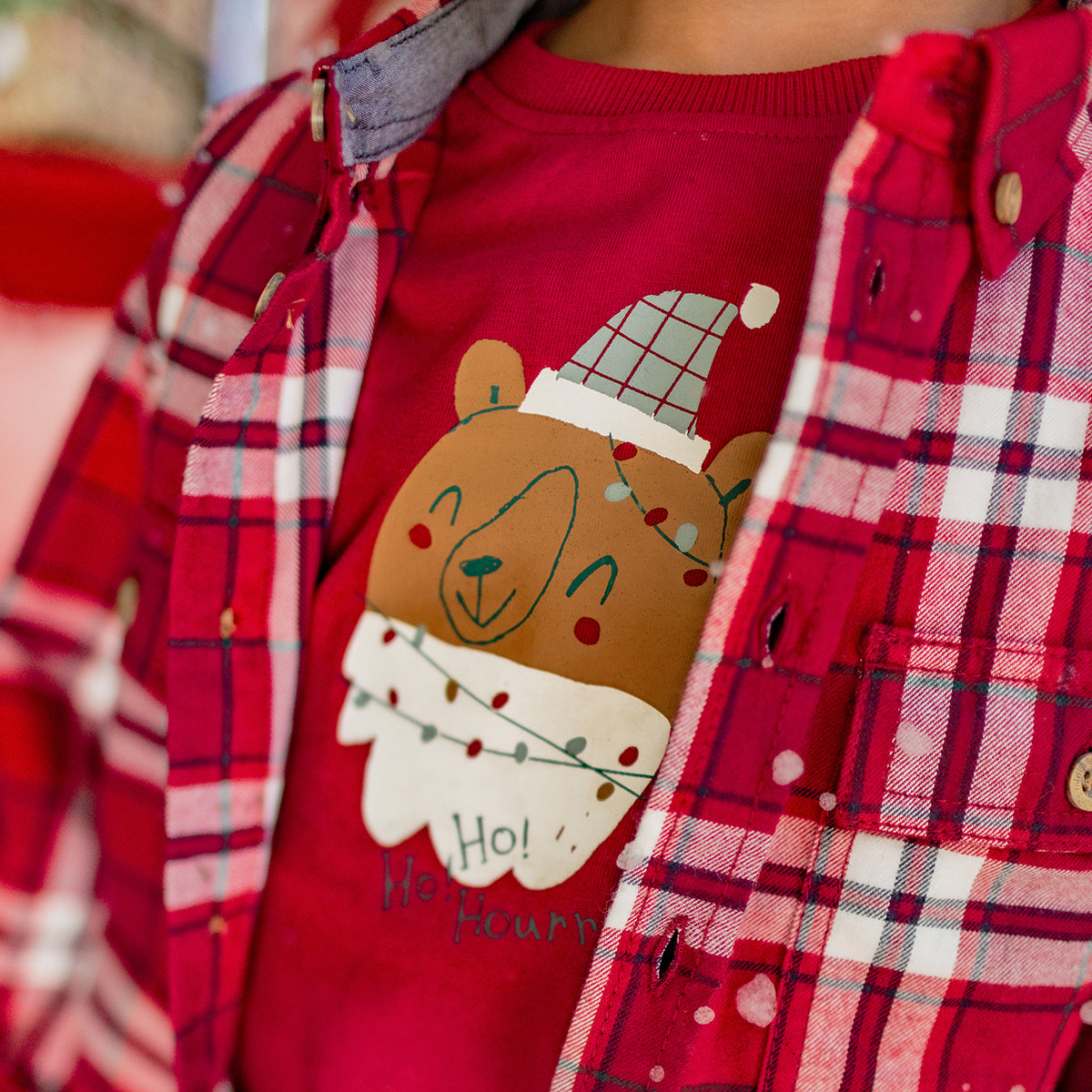 Gros plan sur un t-shirt rouge pour garçon avec illustration d’un ours festif portant une chemise à carreaux rouges, parfait pour Noël.