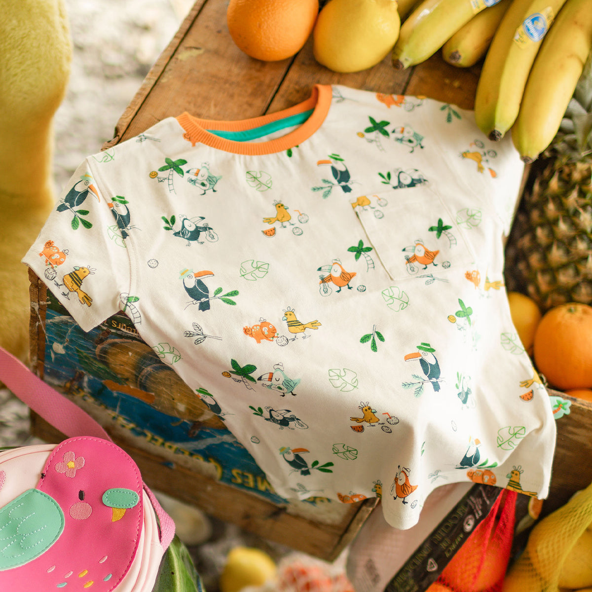 T-shirt crème à motifs tropicaux d’oiseaux et de feuilles, posé sur une caisse en bois parmi des bananes, oranges et autres fruits dans un décor estival.
