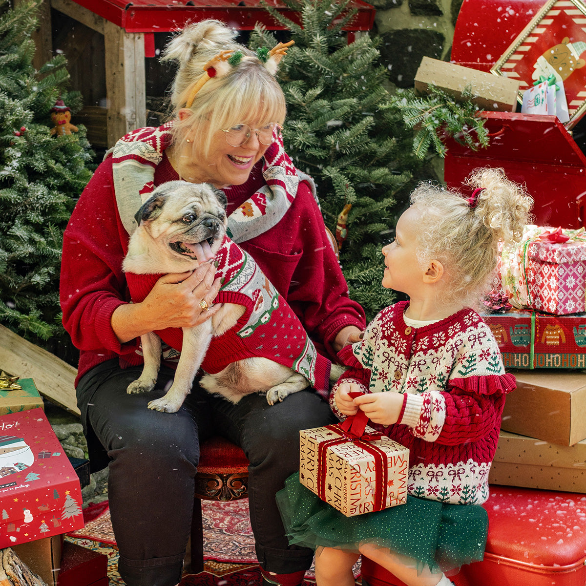 Grand-mère, enfant et chien vêtus de chandails rouges en tricot festifs, partageant un moment de joie autour des cadeaux, collection de Noël Souris Mini.
